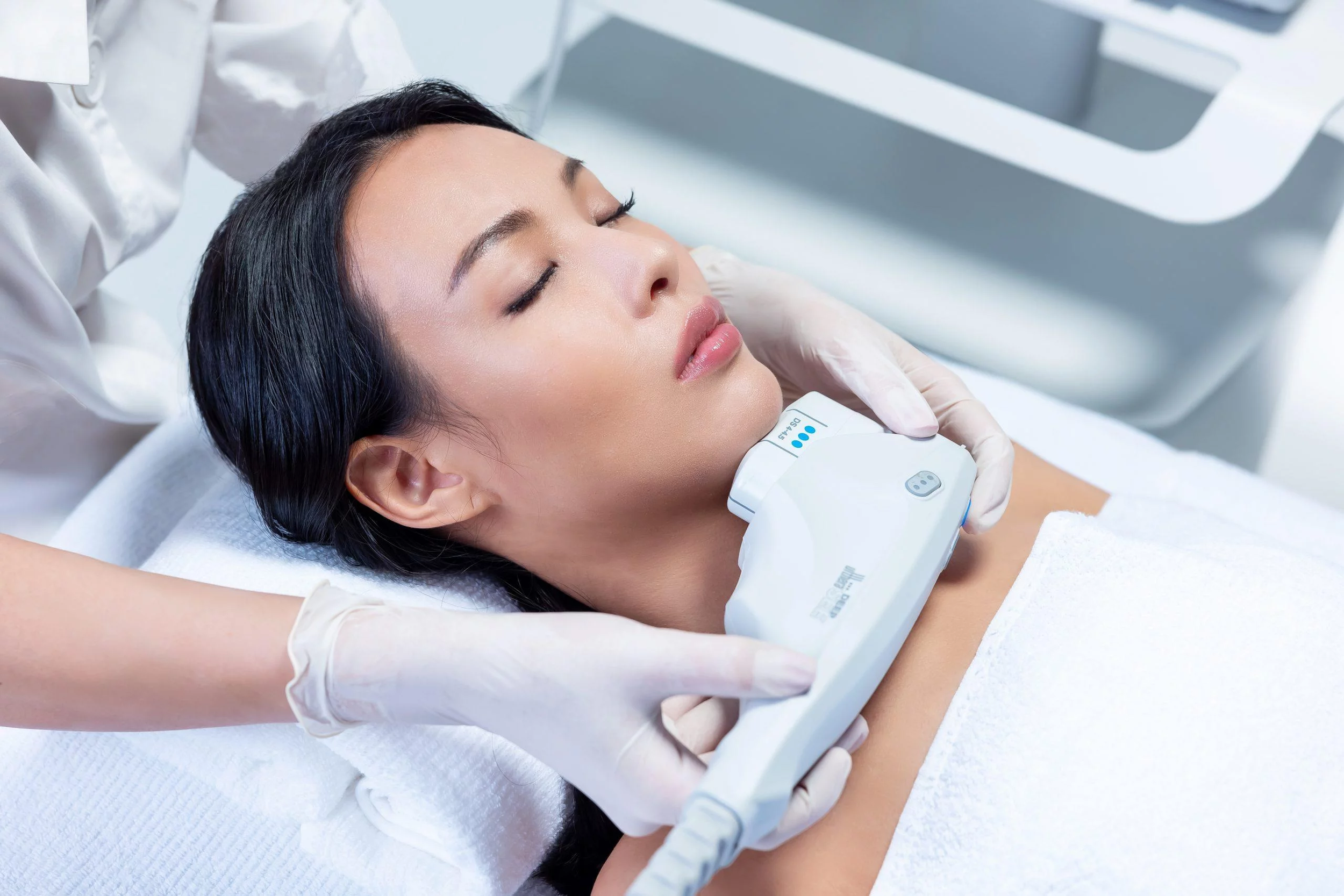 Woman lying down with eyes closed during a facial treatment, receiving a medical skin procedure with a handheld device, in a clinical setting.