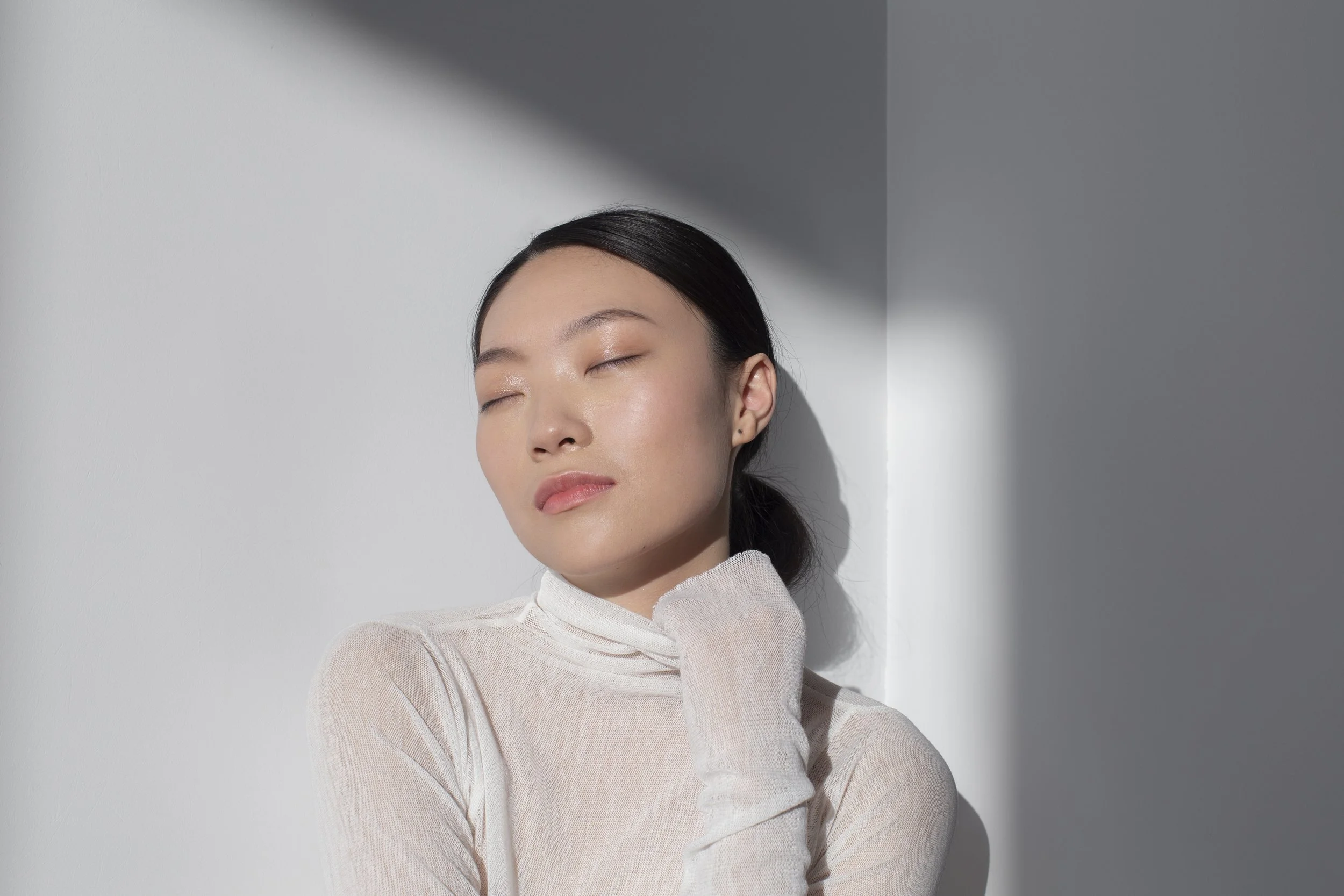 A young woman with dark hair in a ponytail, wearing a sheer, light-colored top, standing with her eyes closed against a white wall, with a shadow cast from the corner.