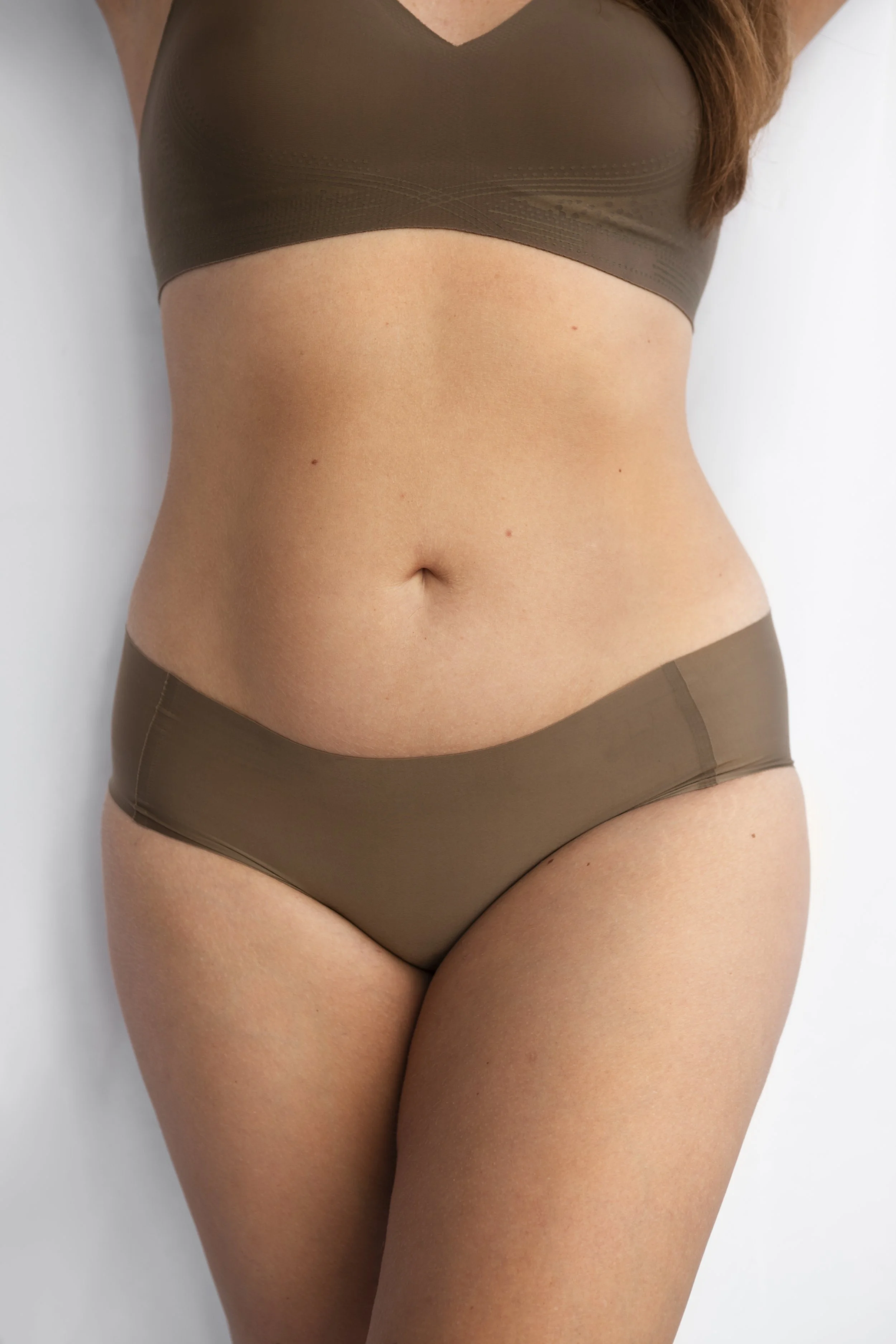 Close-up of a woman's midsection showing her bare abdomen, with her wearing a brown sports bra and matching underwear, standing against a plain, light-colored background.
