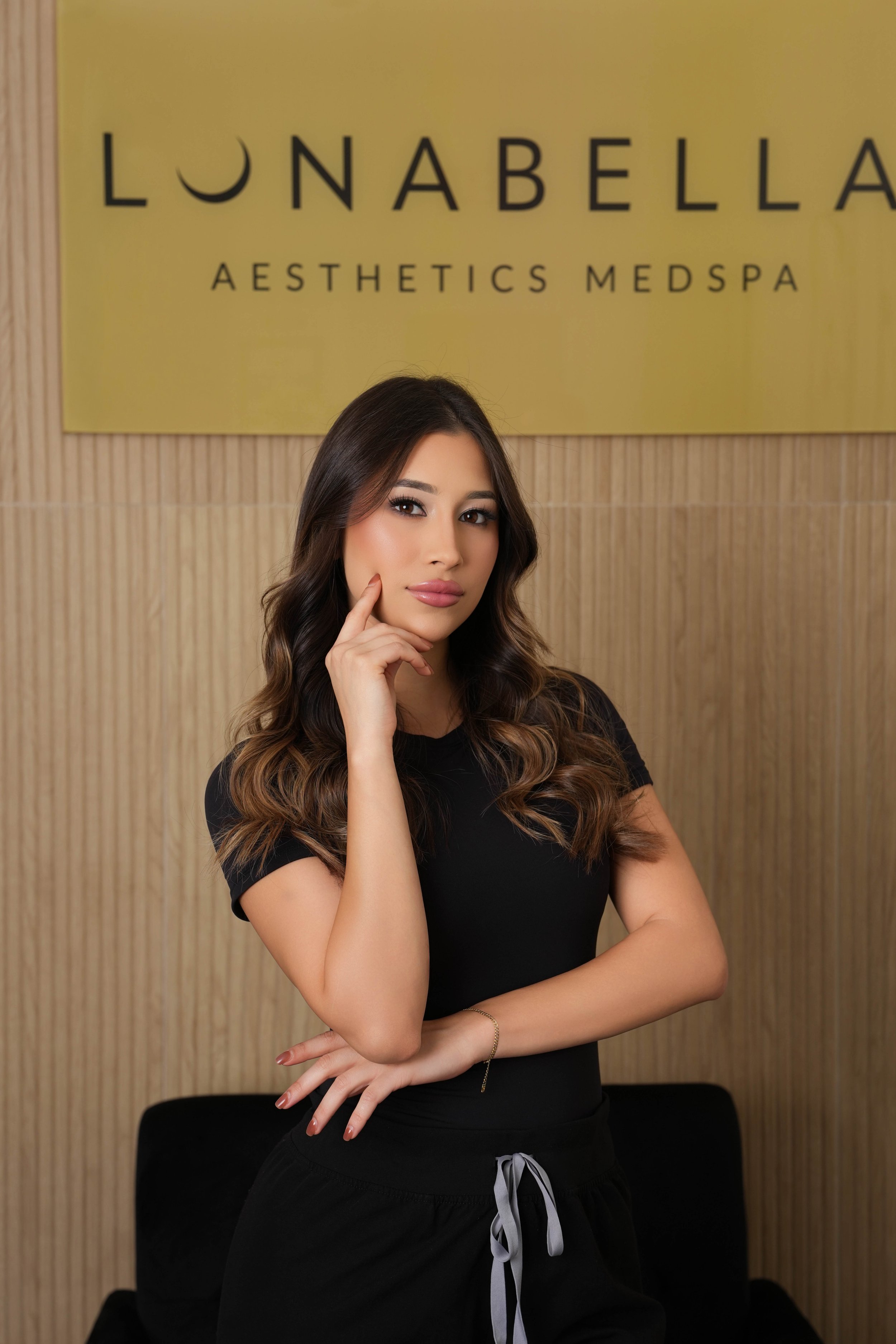 A young woman with long wavy brown hair, wearing a black top and black pants, standing in front of a yellow wall with a sign that reads 'LUNABELLA AESTHETICS MEDSPA', posing with her hand on her chin and a confident expression.