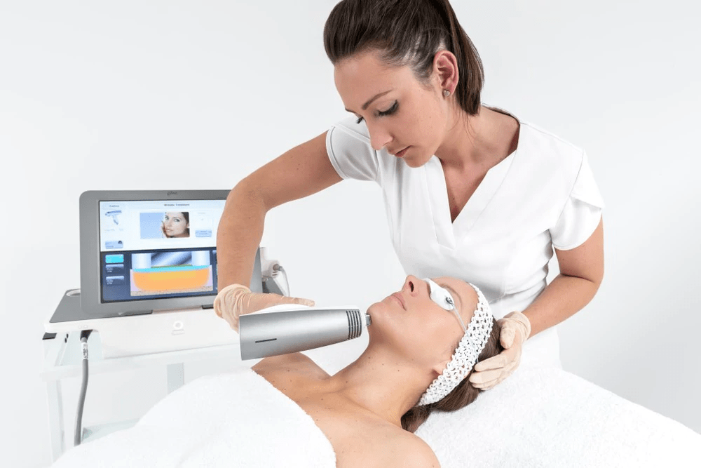 A woman receiving a facial treatment from an aesthetic professional, lying on a treatment bed with a headband on, in a clinical setting.