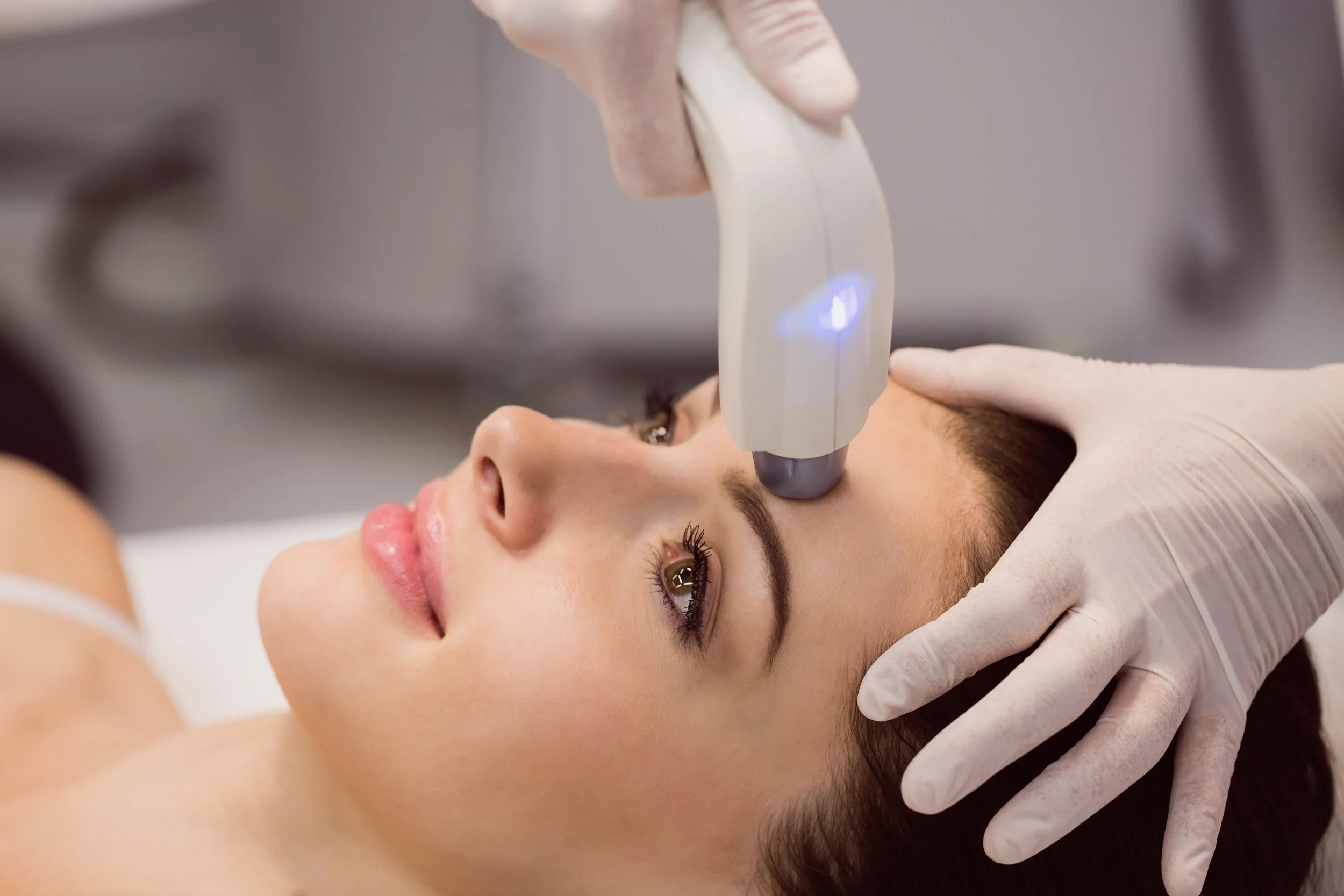 A woman is lying down during a cosmetic treatment, with a technician in gloves applying a device to her face.