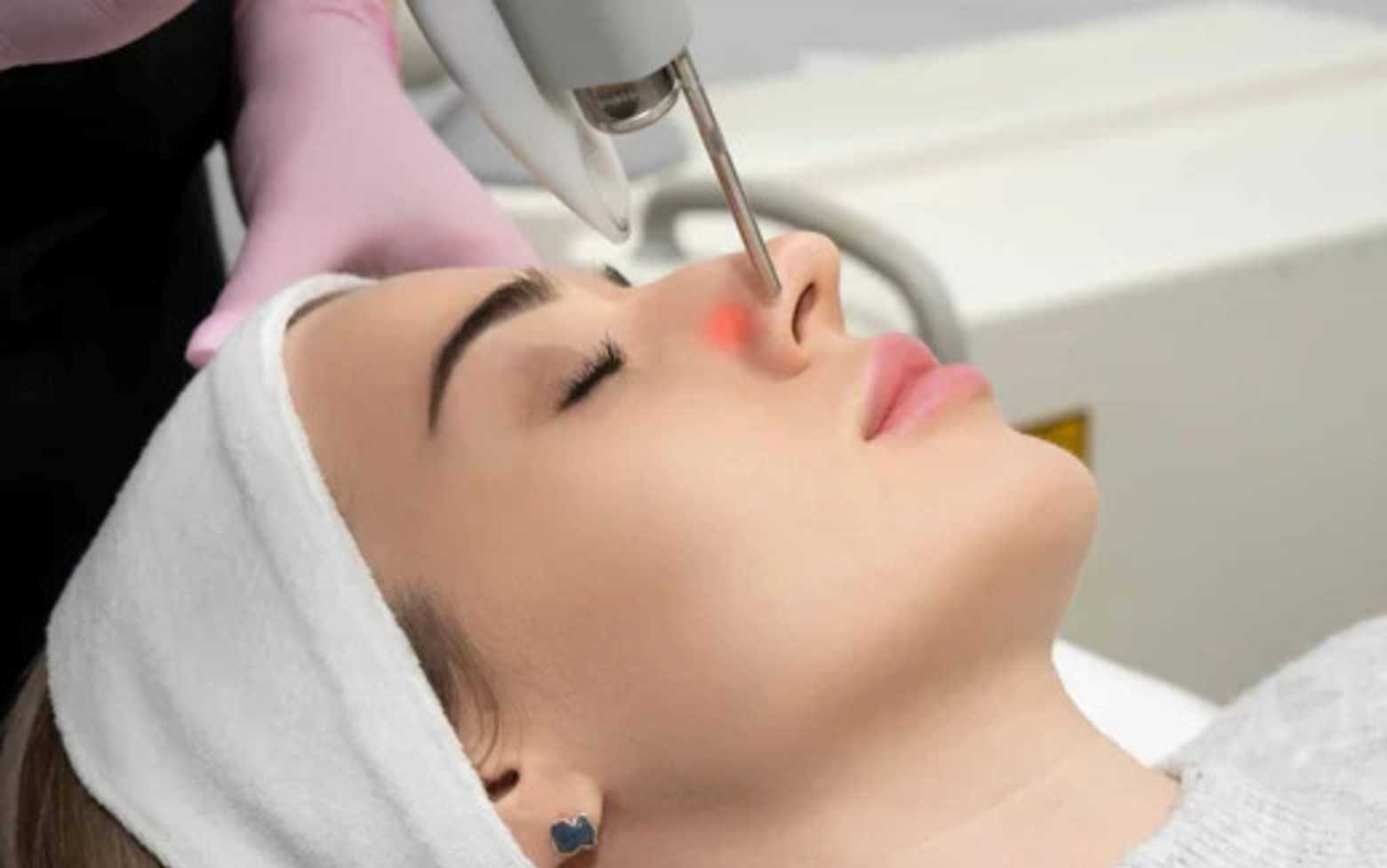 A woman receiving a non-invasive facial treatment with a specialized device on her nose at a clinic.