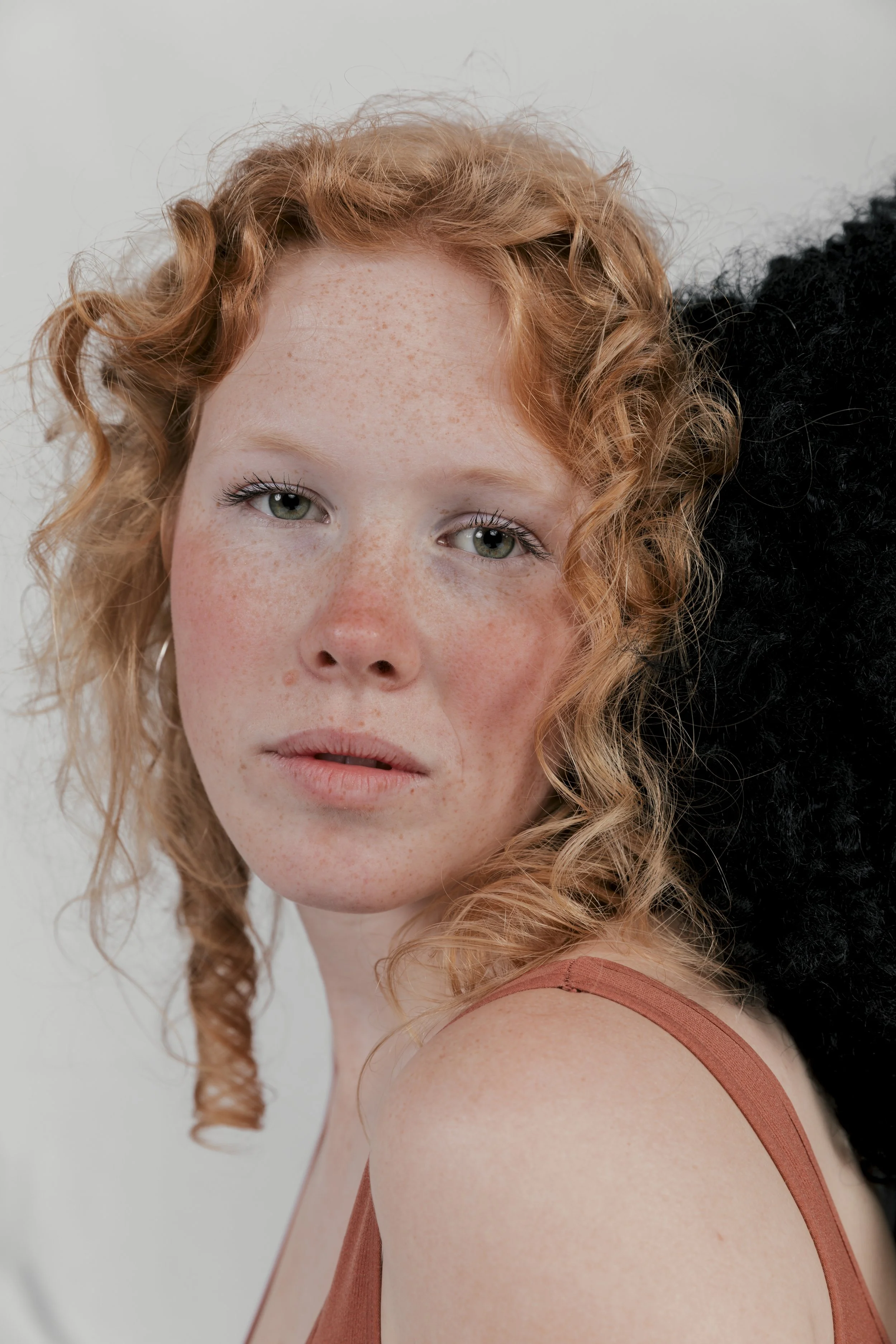 Close-up portrait of a young woman with red curly hair, green eyes, freckles, and wearing a sleeveless rust-colored top, against a plain light background.