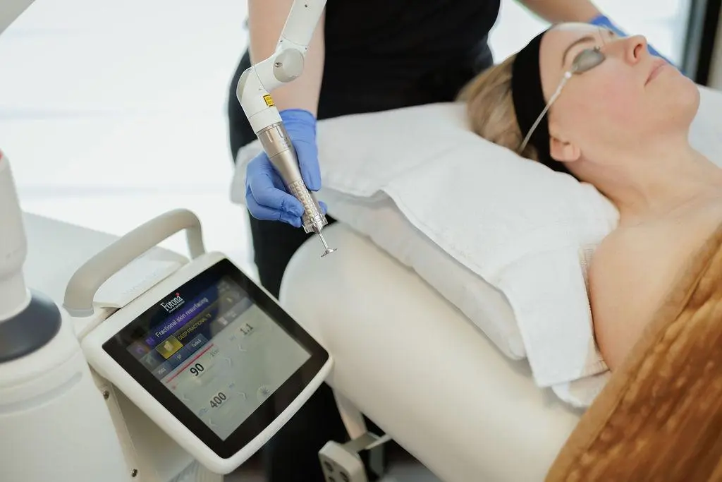 A woman with blonde hair and earrings lying on a treatment bed. She is wearing protective goggles and receiving a facial treatment with a handheld device.