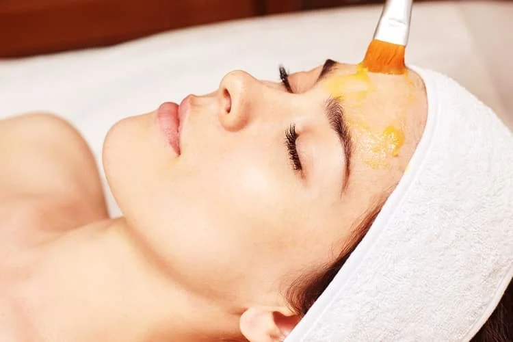 A woman receiving a facial treatment, with a brush applying a yellow mask or cream to her forehead while she lies peacefully with her eyes closed.