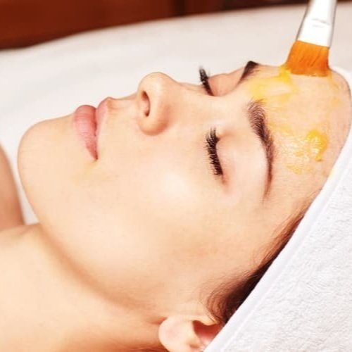 A woman lying down with her eyes closed, receiving a facial treatment with a brush applying a yellow cream or mask on her forehead.