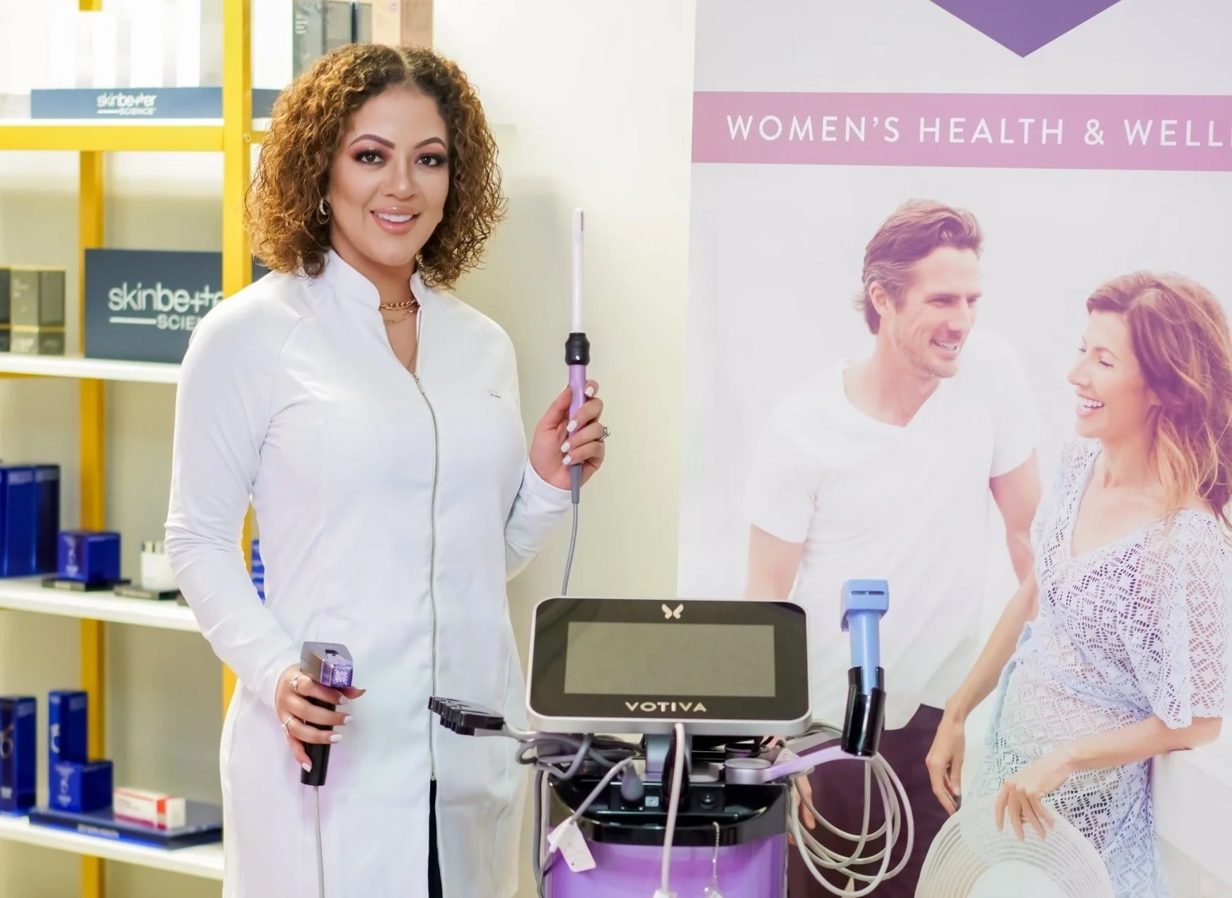 A woman with curly hair smiling while holding a medical device in a healthcare setting, with a banner in the background that reads 'Women's Health & Wellness' and a poster of a happy couple.