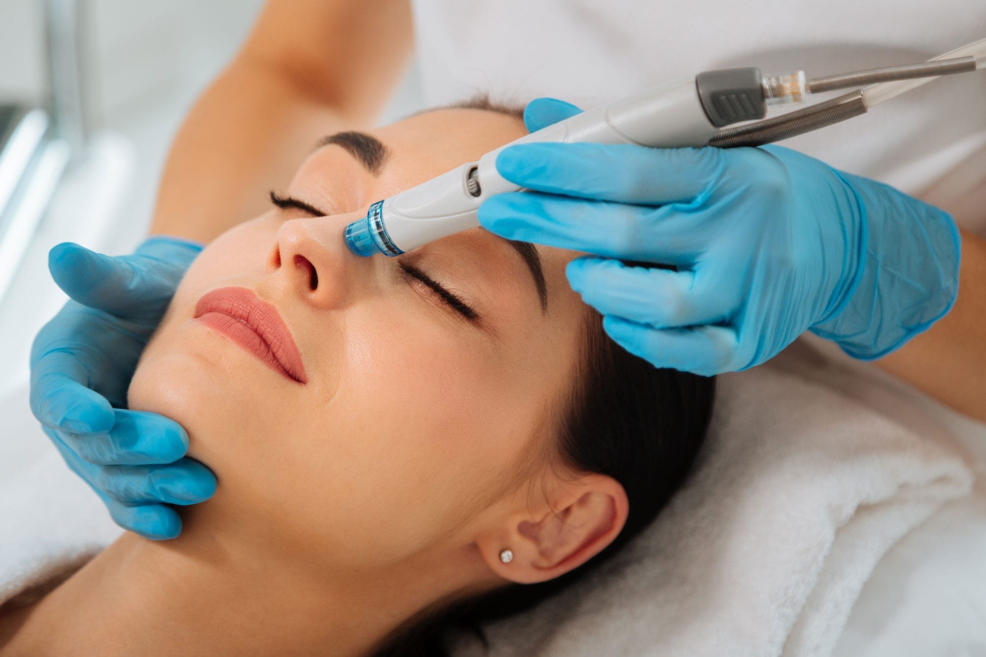 A woman receiving a facial treatment with a handheld device from a professional wearing blue gloves.