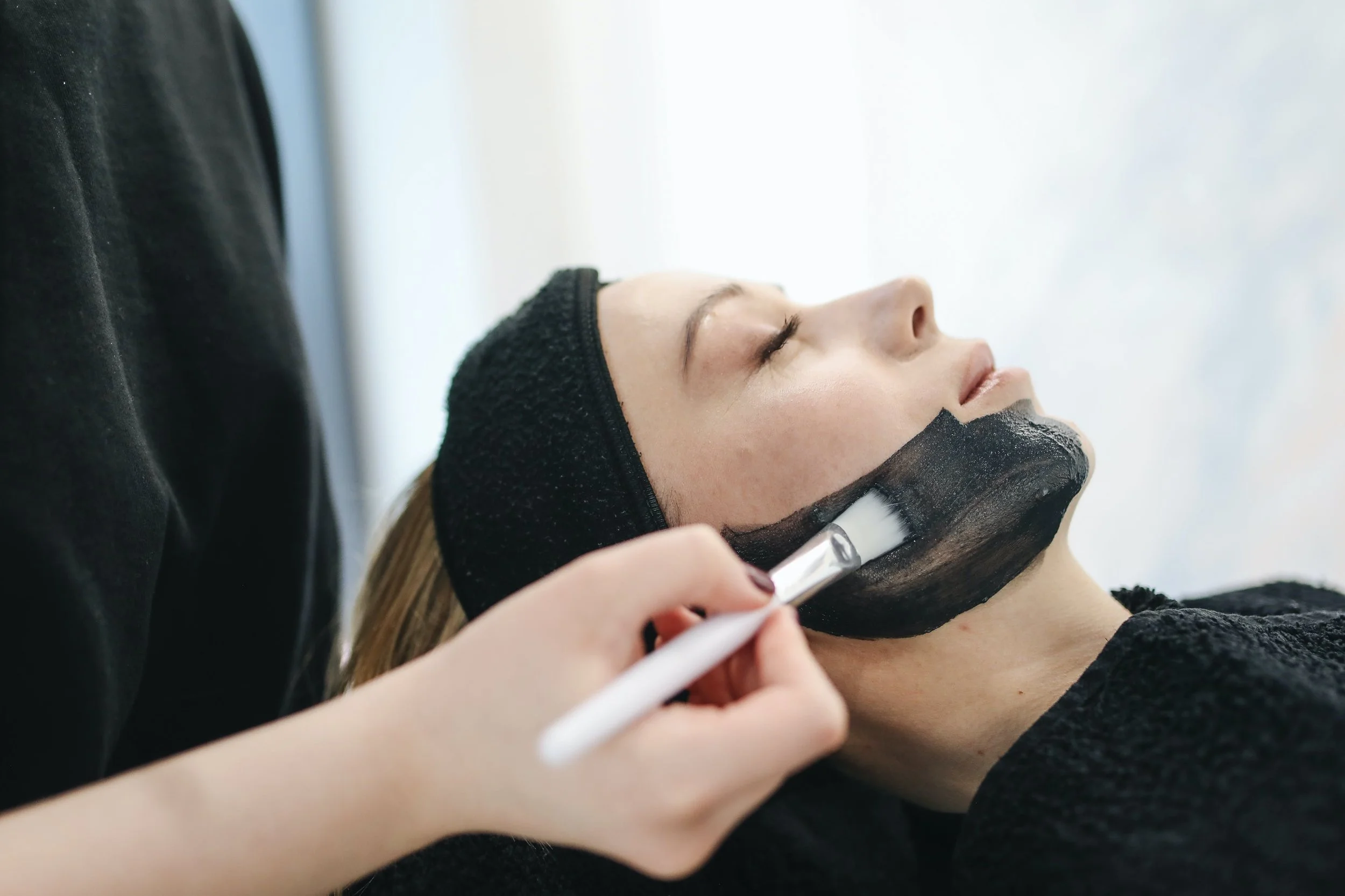 A woman is receiving a facial treatment; she has her eyes closed and is lying down while a esthetician applies a black facial mask with a brush.