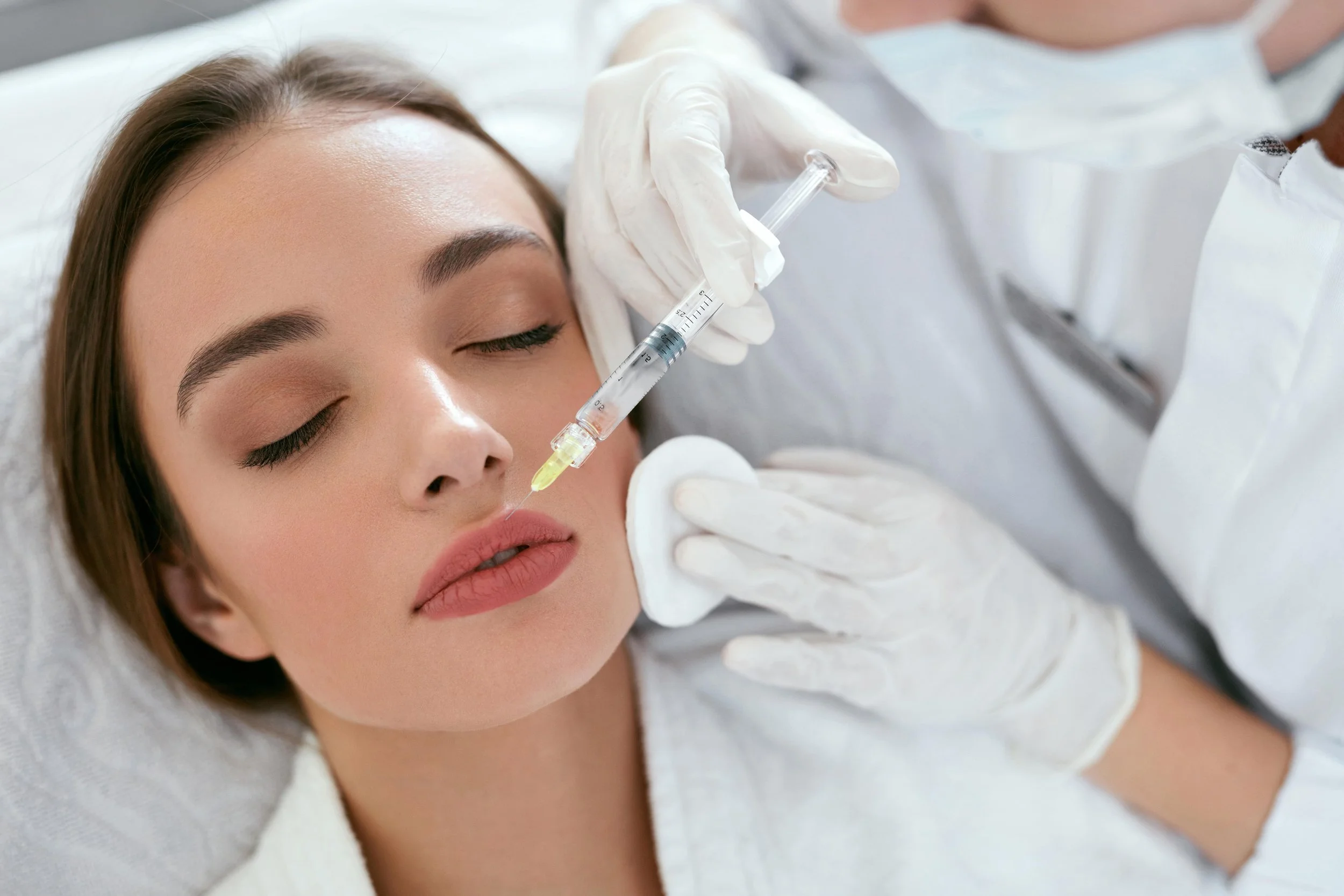 A woman with closed eyes receiving a cosmetic or medical injection into her face from a healthcare professional wearing gloves.