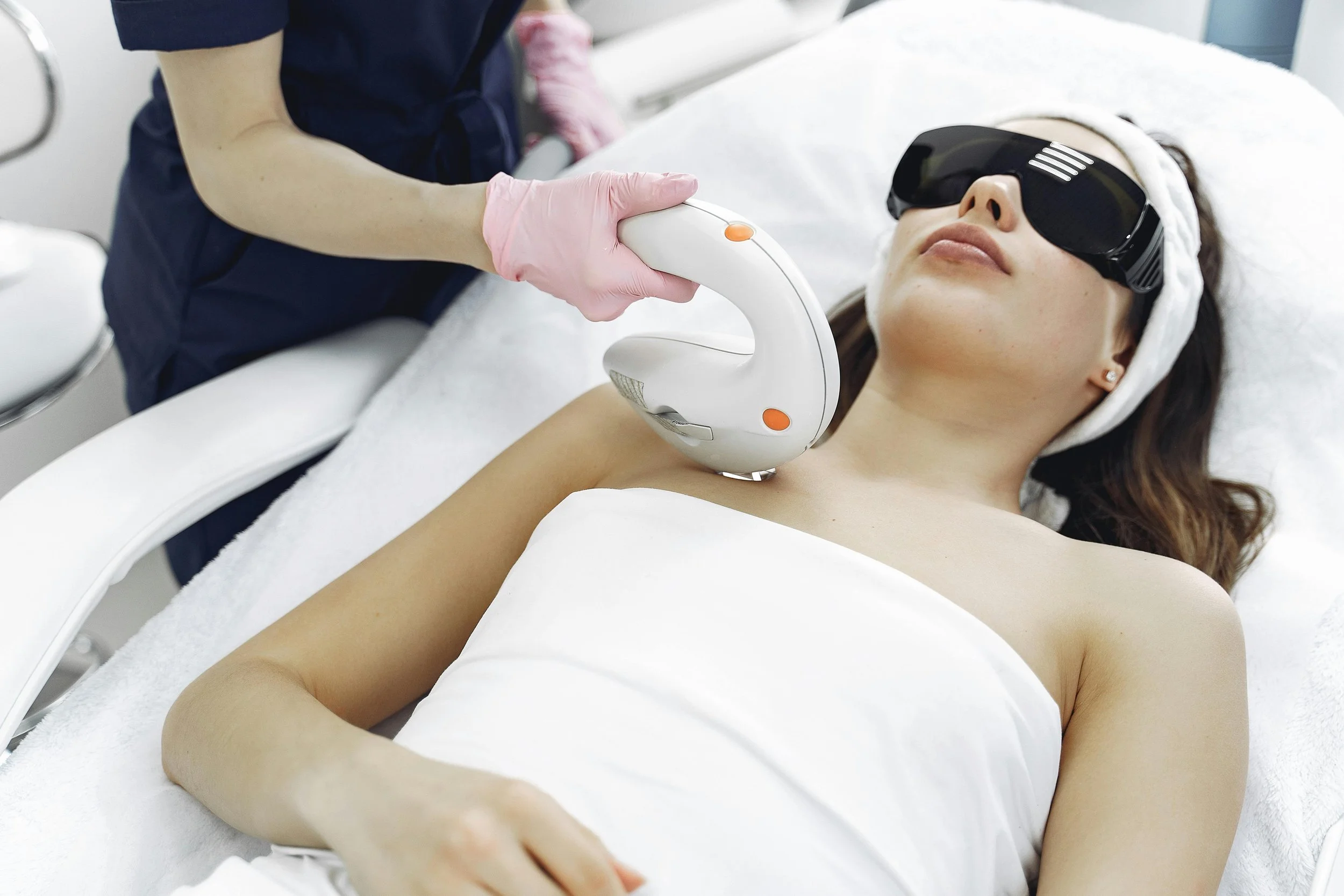 A woman lying on a treatment bed wearing protective glasses, while a person in medical gloves uses a handheld device on her chest for a cosmetic or medical procedure.