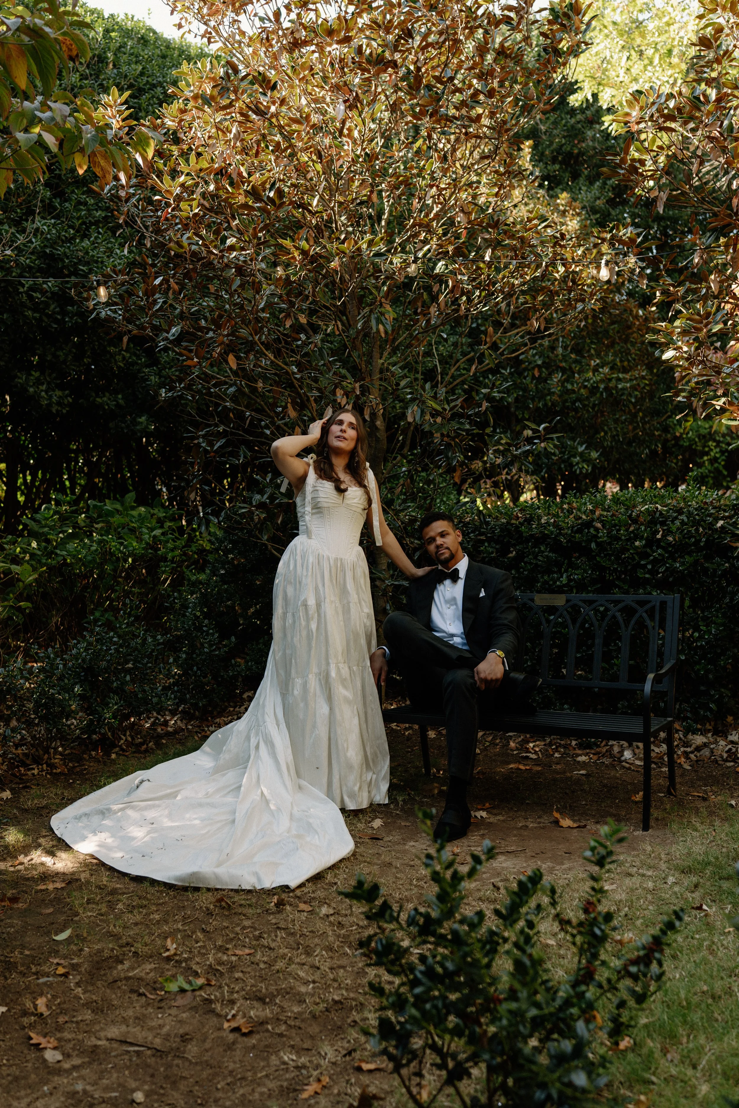 A woman in a long, white wedding dress and a man in a black tuxedo sit on a black metal park bench in a lush garden. The woman is standing and touching her hair, while the man is sitting with one leg crossed, resting his arm on the back of the bench.