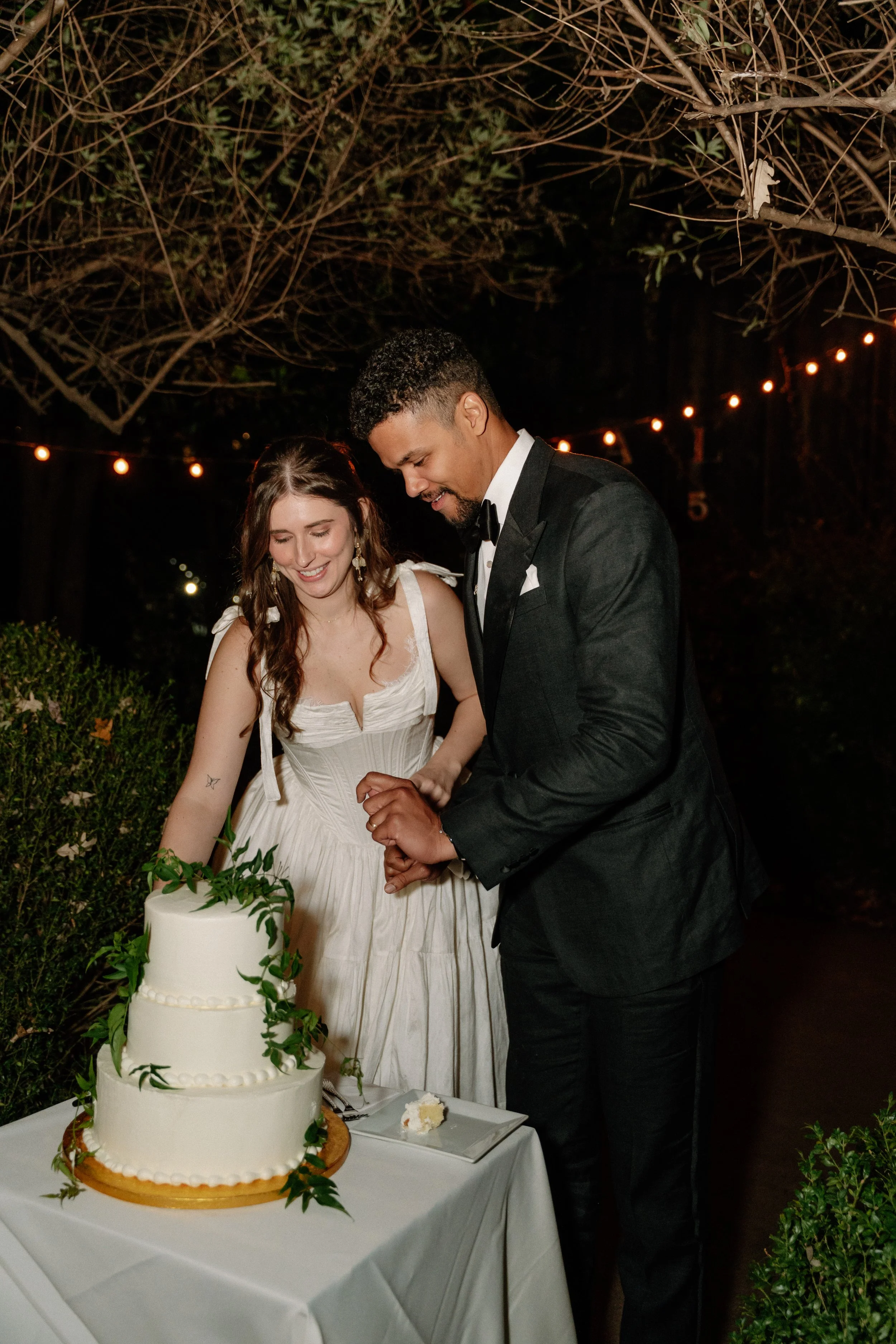 A bride and groom cutting their wedding cake outdoors at night, decorated with green leaves and string lights above.