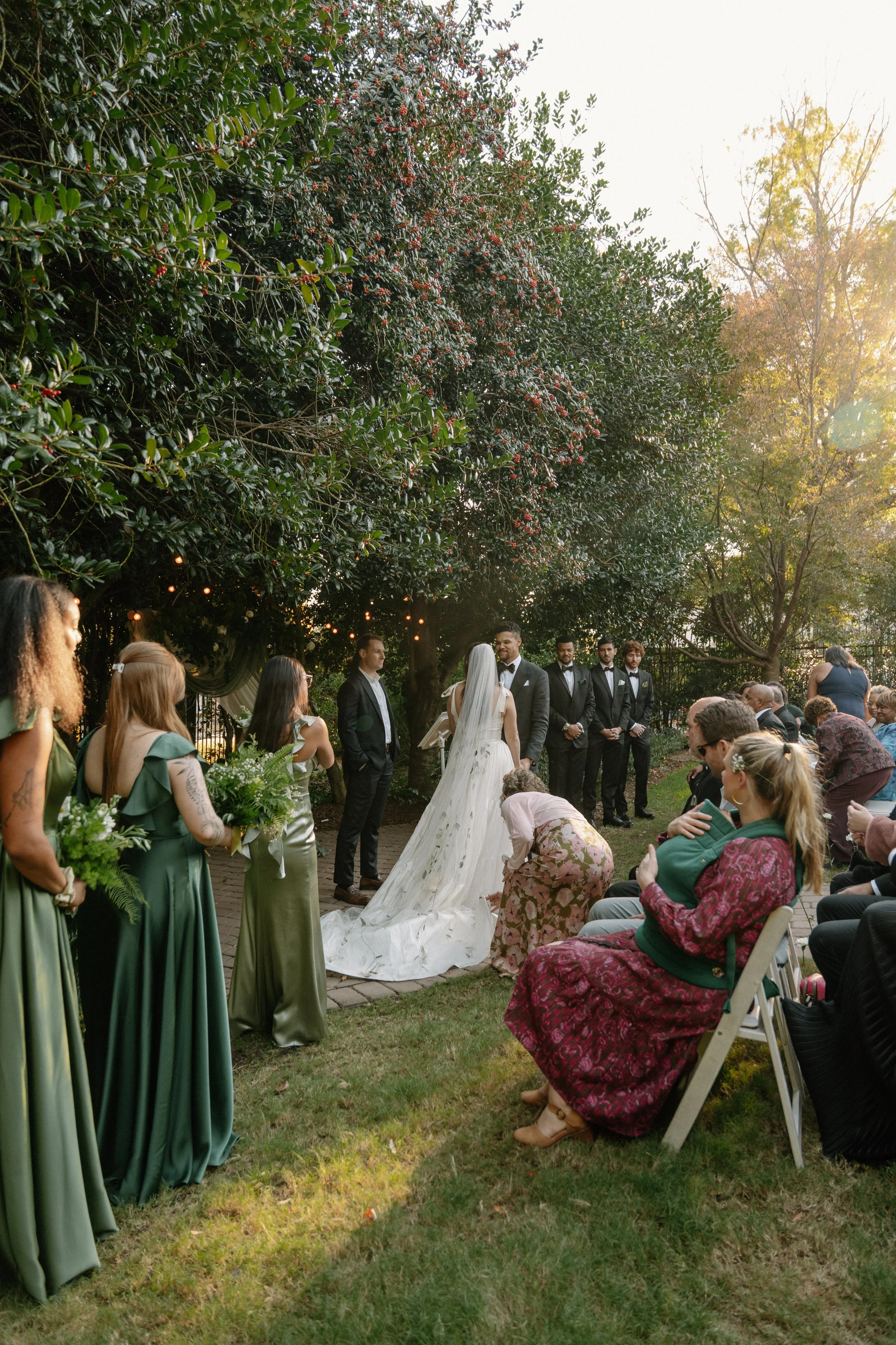 A wedding ceremony taking place outdoors under trees with sunlight filtering through. The bride and groom stand facing each other, surrounded by bridesmaids and groomsmen. Guests are seated, watching the ceremony, with some taking photos. The scene i