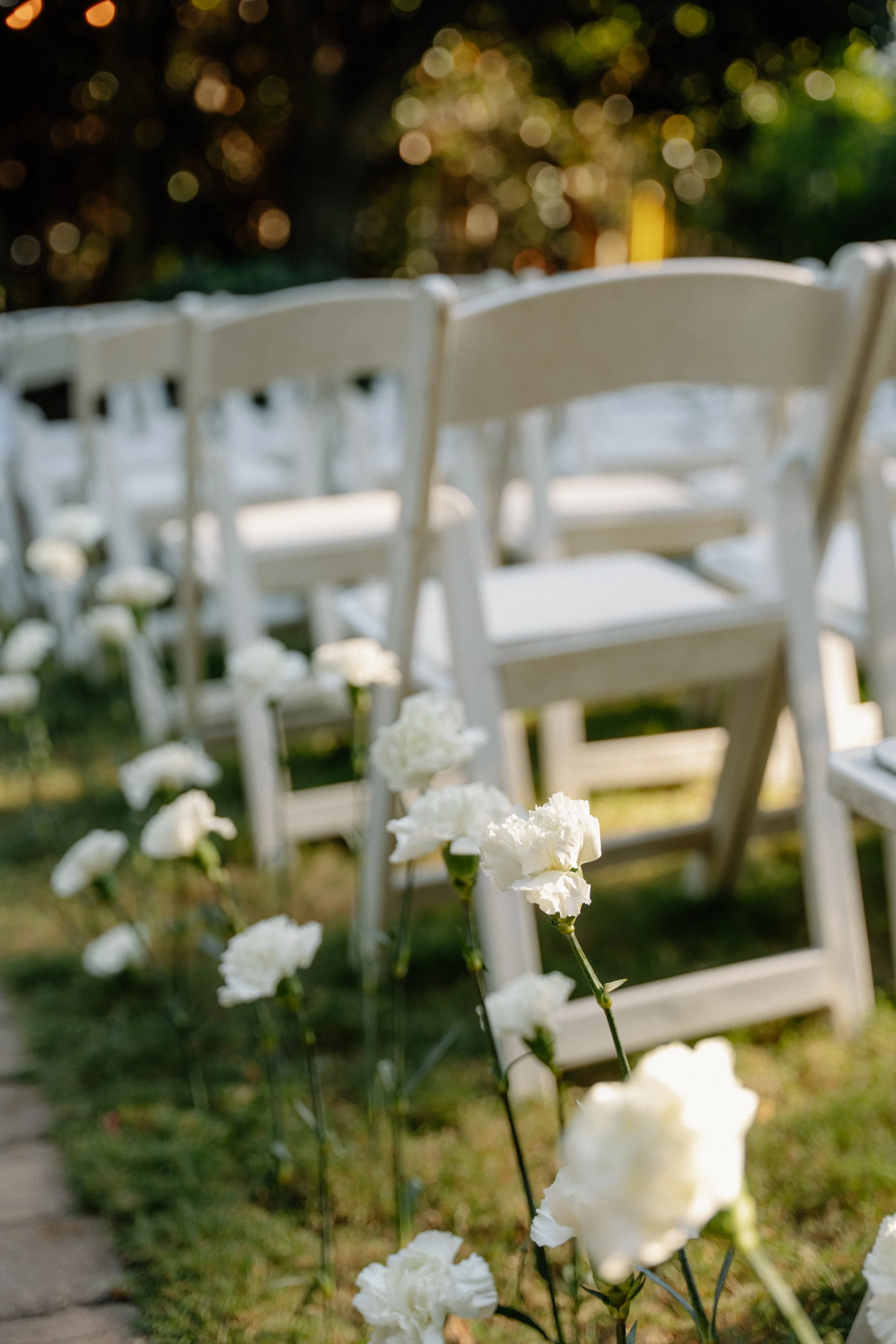 White chairs arranged outdoors with white flowers lining the path at a wedding ceremony.