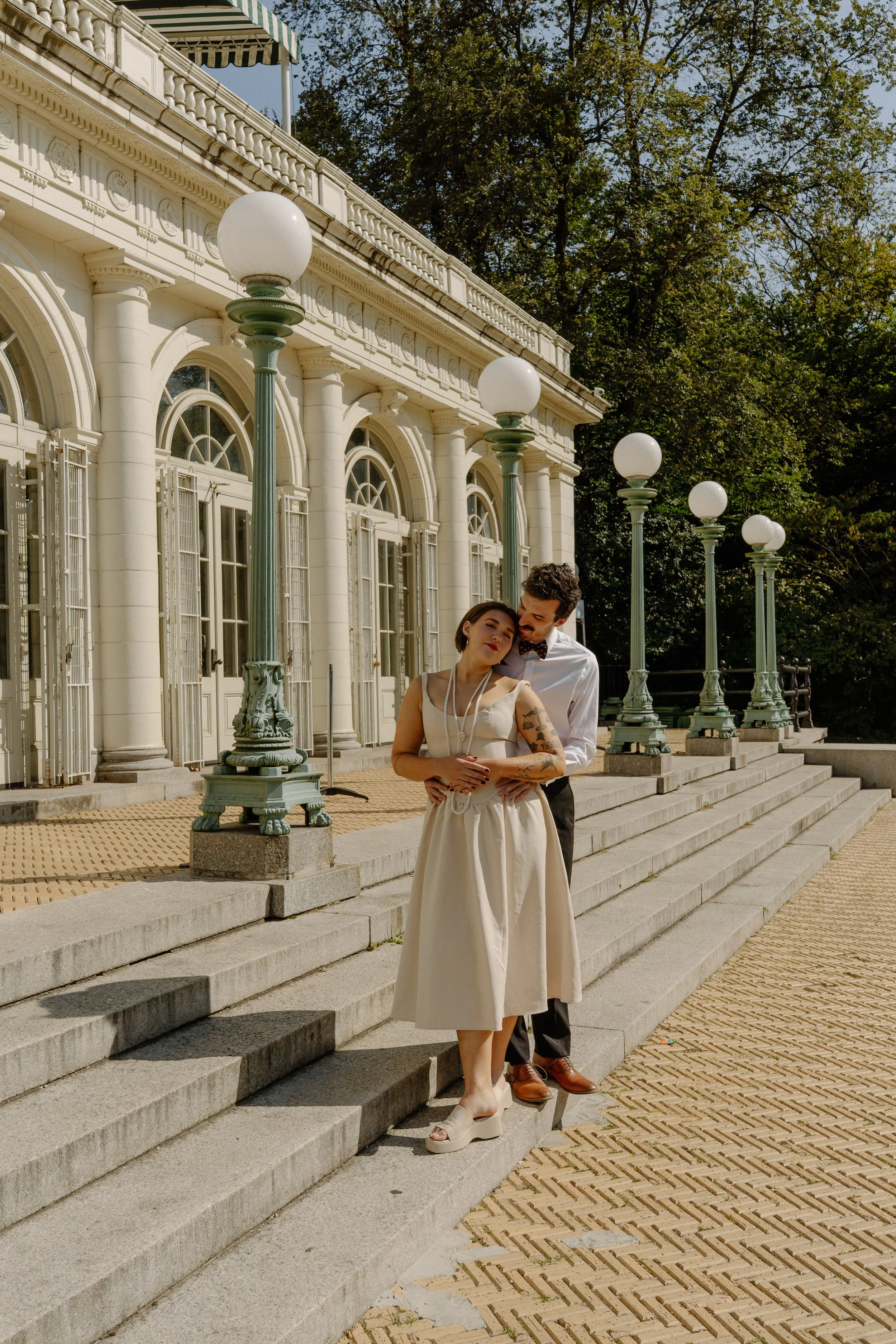 A couple stands on the steps of a historic building with arched windows and columns, surrounded by ornate street lamps. The woman is dressed in a vintage cream dress with platform shoes and has tattoos. The man is dressed in a white shirt, black pant