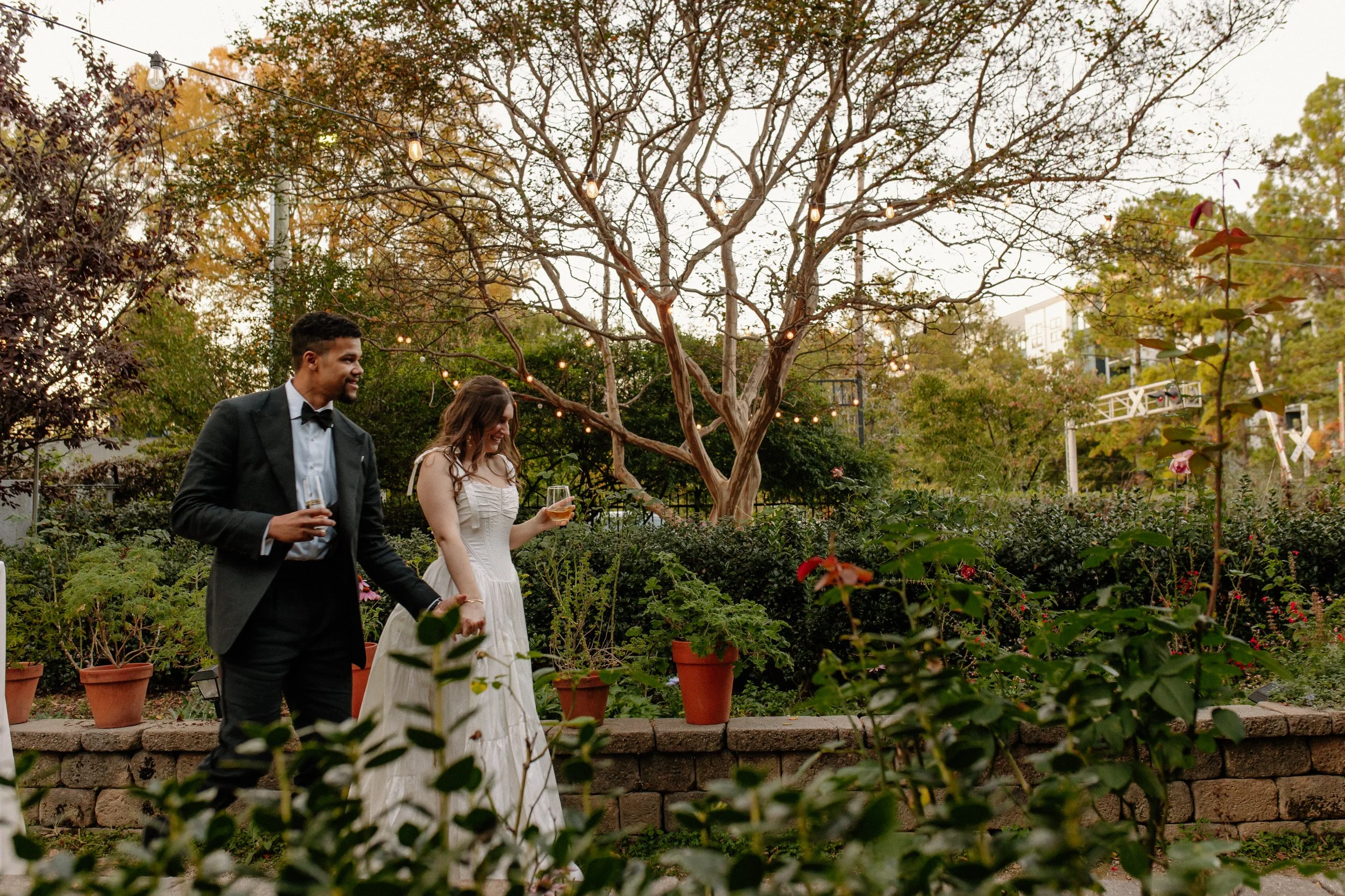 A couple dressed in formal attire, holding glasses of champagne, walking outdoors in a garden with potted plants, trees, and string lights during the evening.
