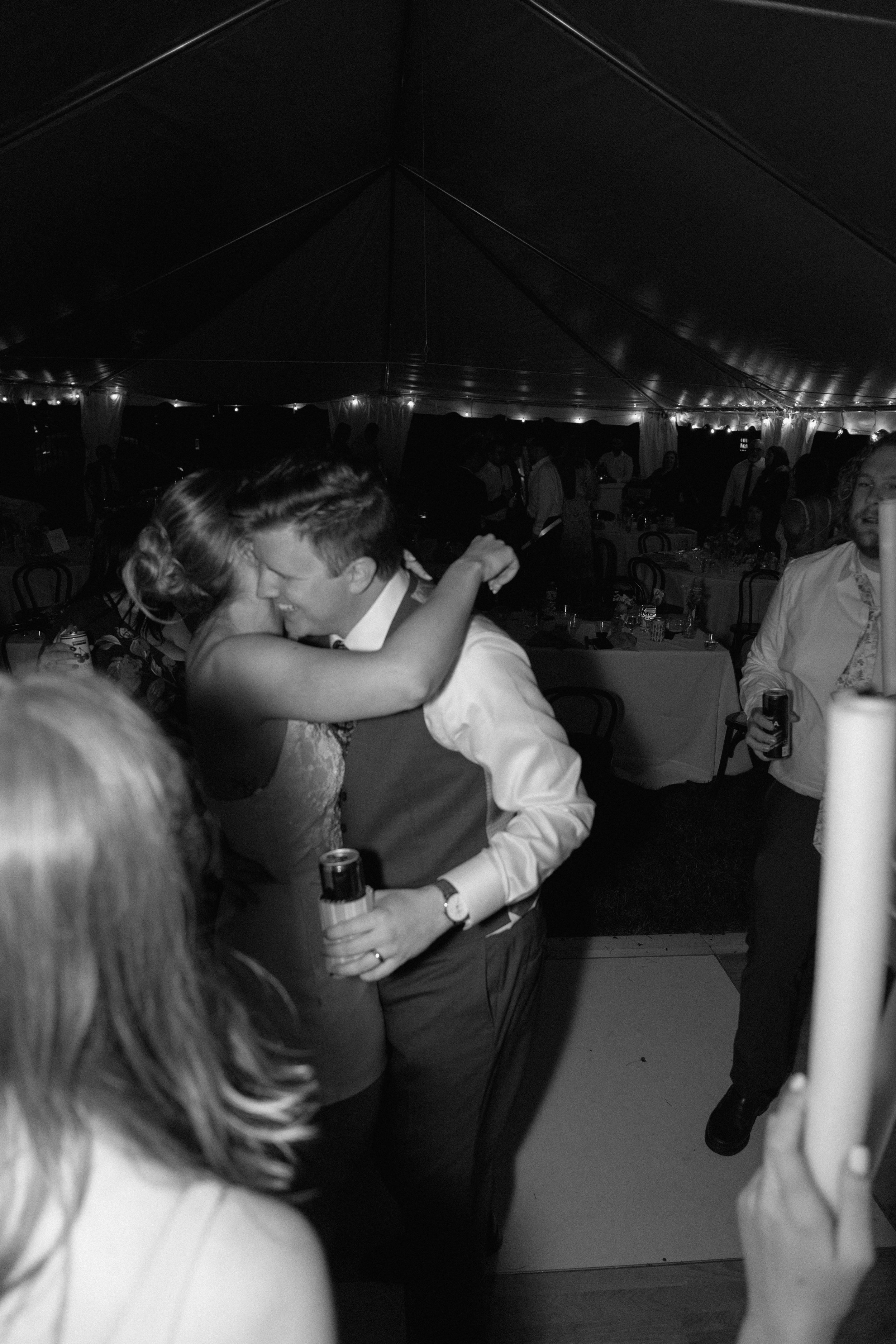 A black-and-white photo of a couple kissing at a wedding reception inside a large tent, surrounded by guests and decorated tables.