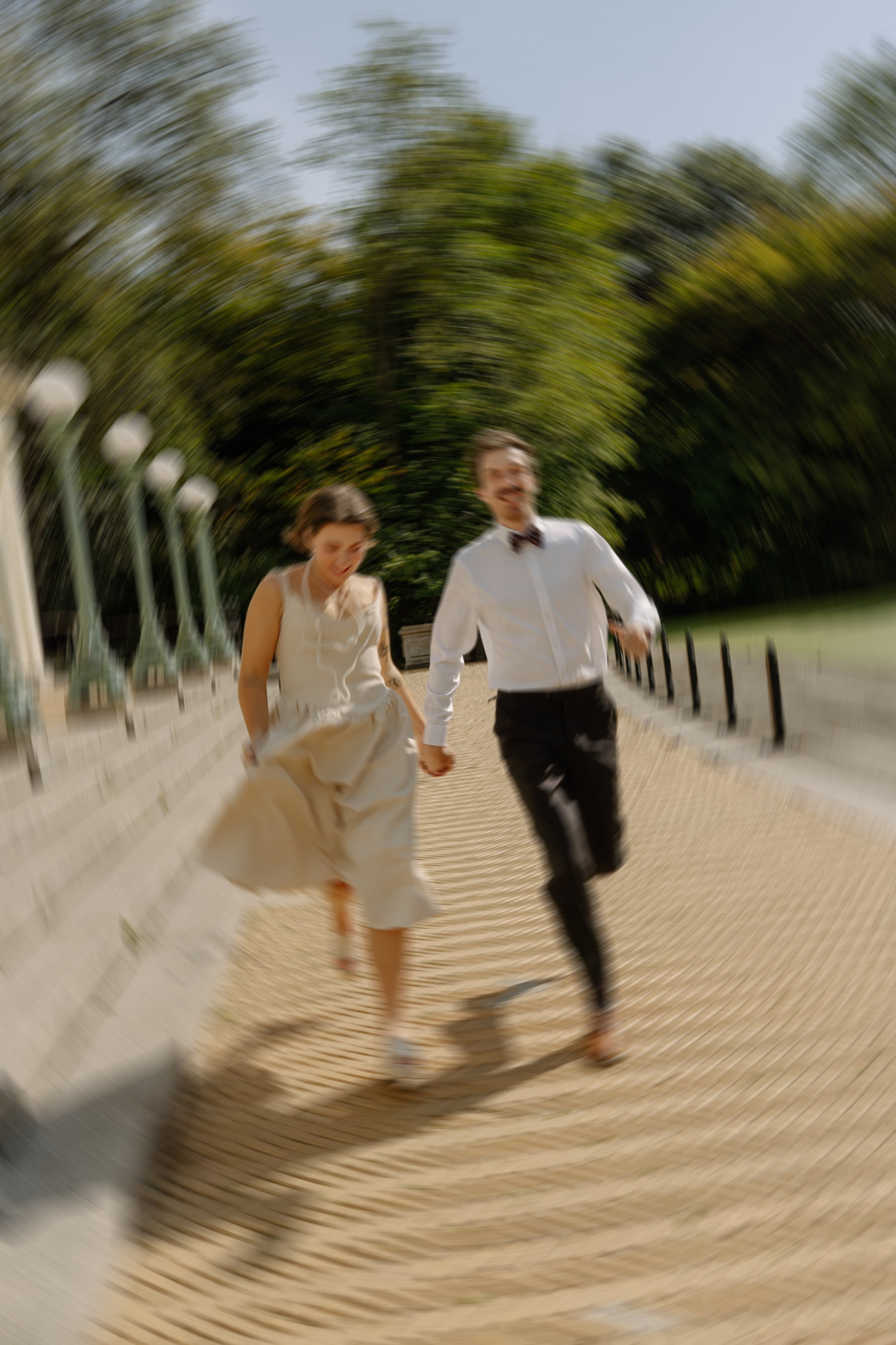 A couple holding hands and running barefoot along a paved pathway in a park with green trees, captured with a motion blur effect.