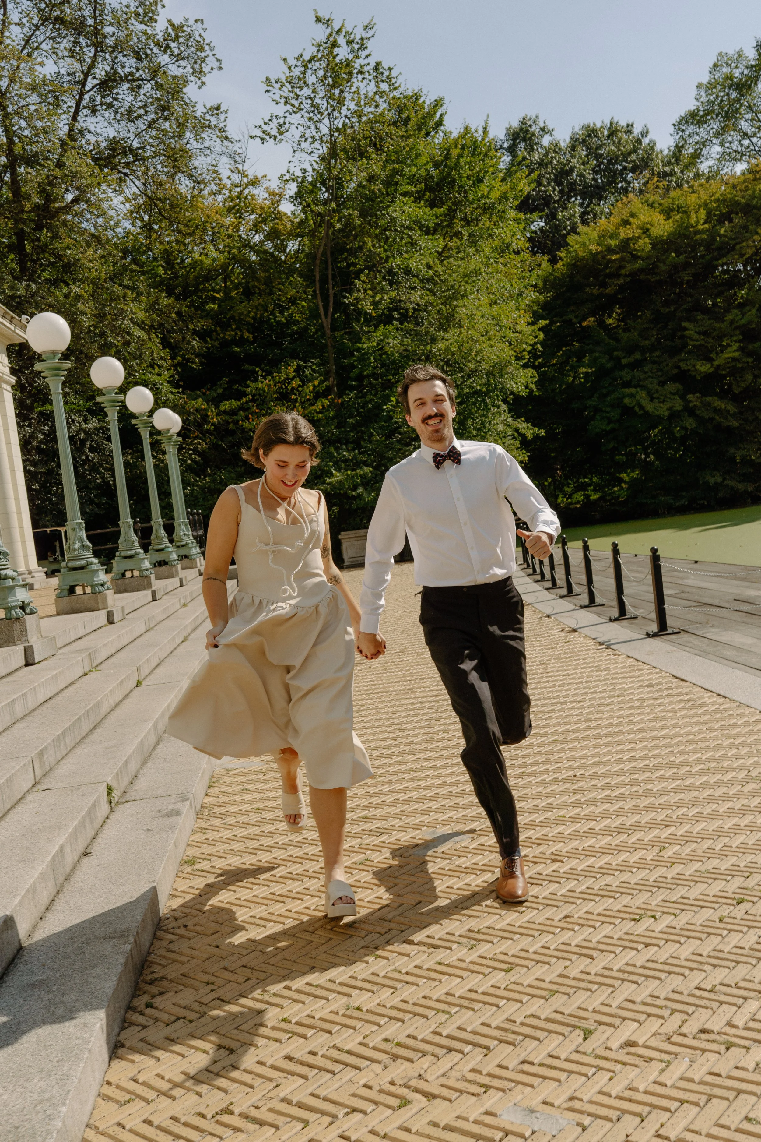 A couple, a woman in a beige dress and a man in a white shirt with a bow tie, is holding hands and running joyfully on a brick pathway outside amid lush green trees on a sunny day.