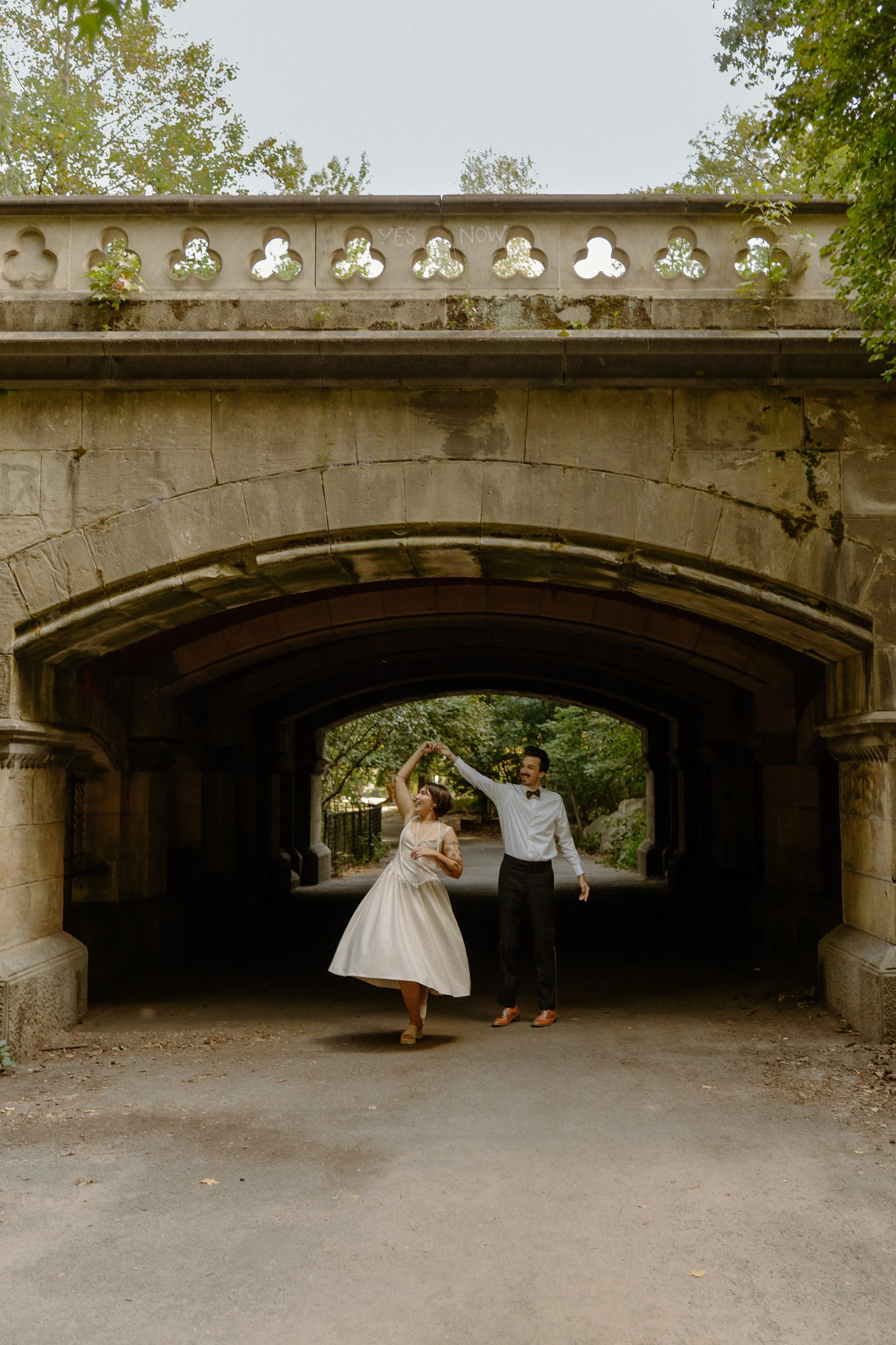 A couple dancing under a stone bridge in a park, with the woman in a vintage dress and the man in formal attire, surrounded by greenery.