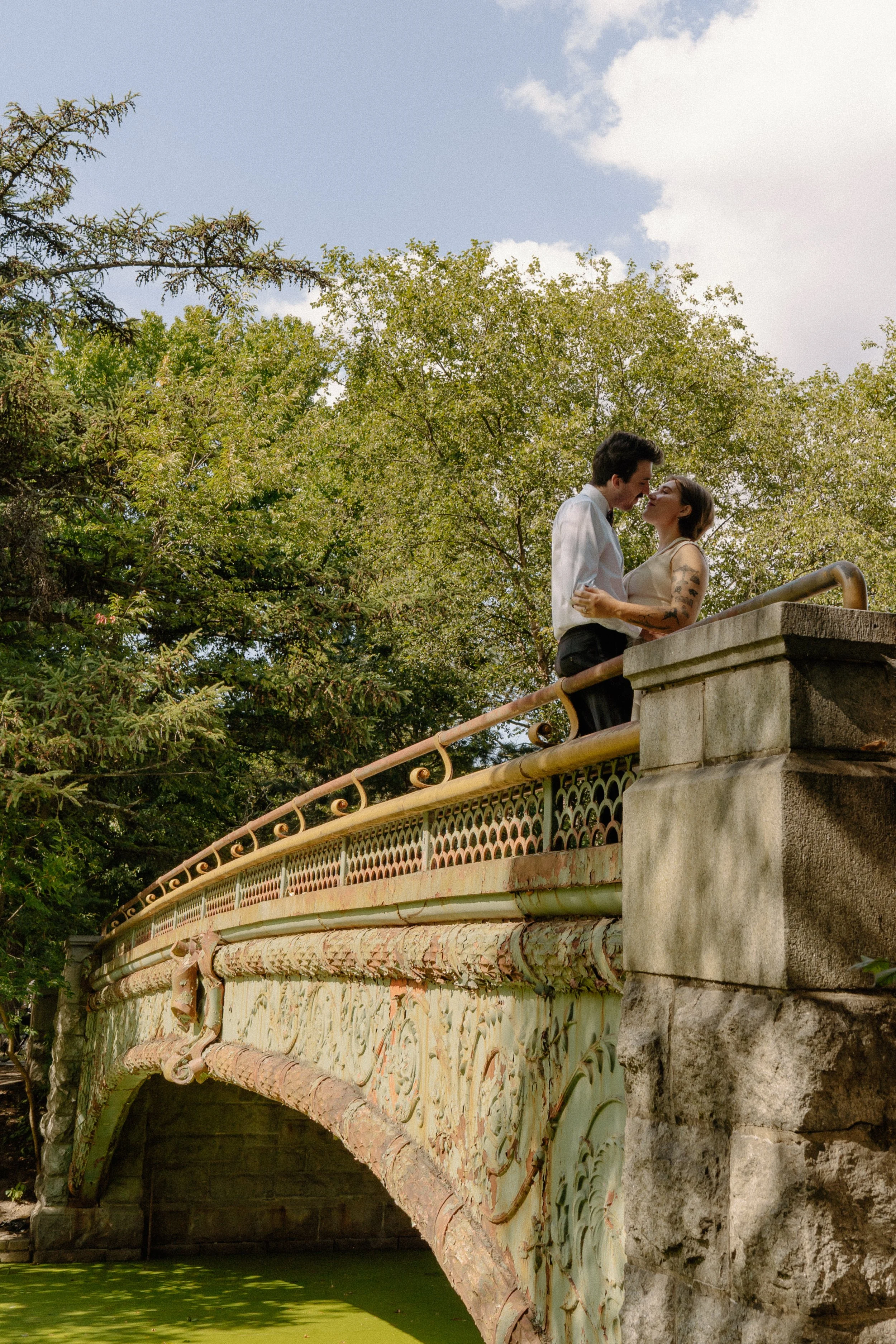 A couple standing close together on a decorative old bridge, embracing and about to kiss, surrounded by green trees and a partly cloudy sky.