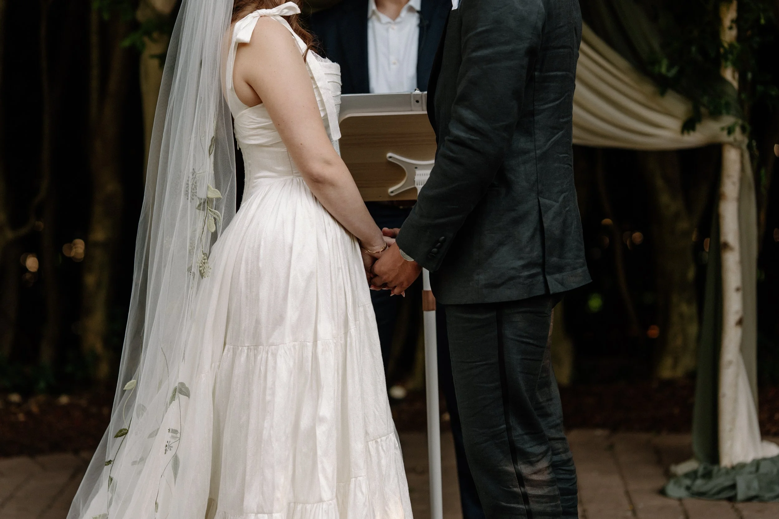 A bride and groom holding hands during their wedding ceremony, with a officiant in the background, outdoors.