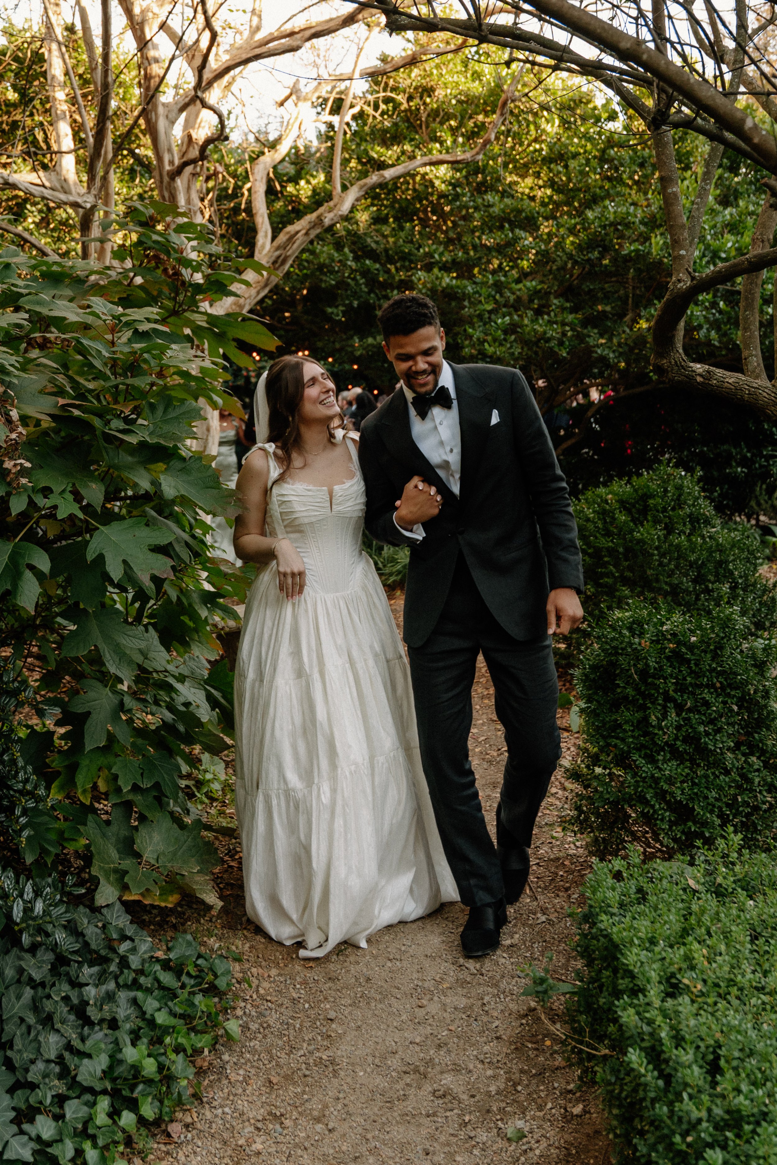 A bride and groom walking arm in arm on a garden path during their wedding, surrounded by trees and greenery, smiling and enjoying each other's company.