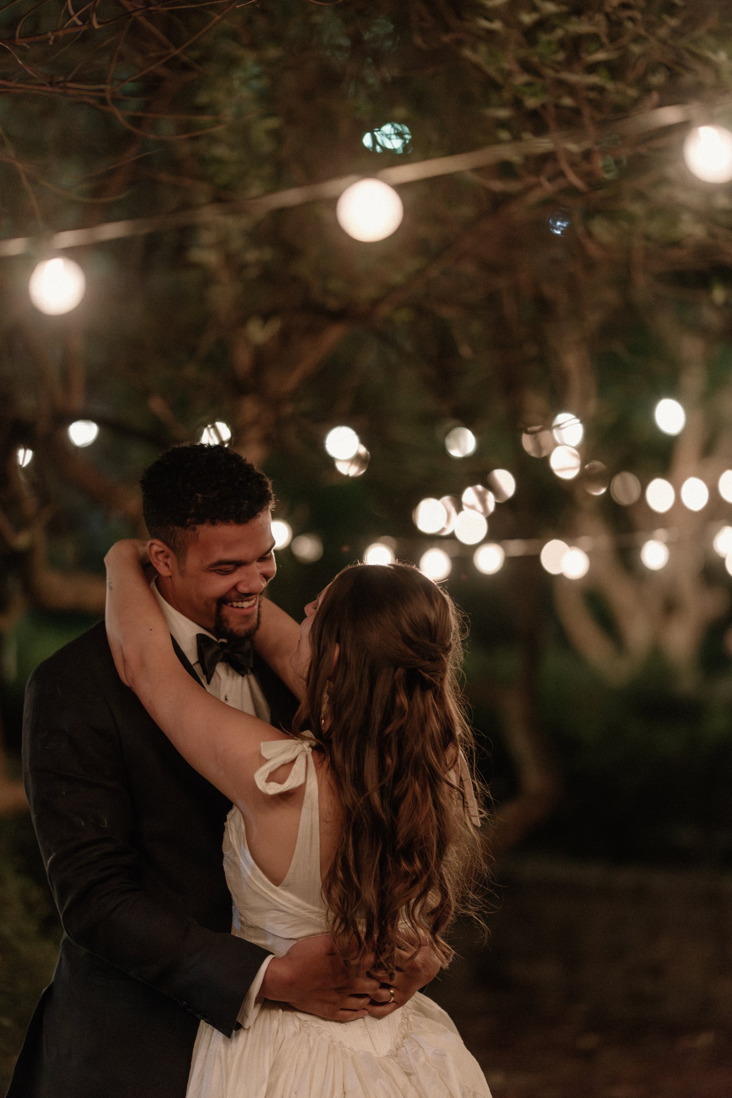 A couple dancing and smiling at night, with string lights hanging from tree branches in the background.