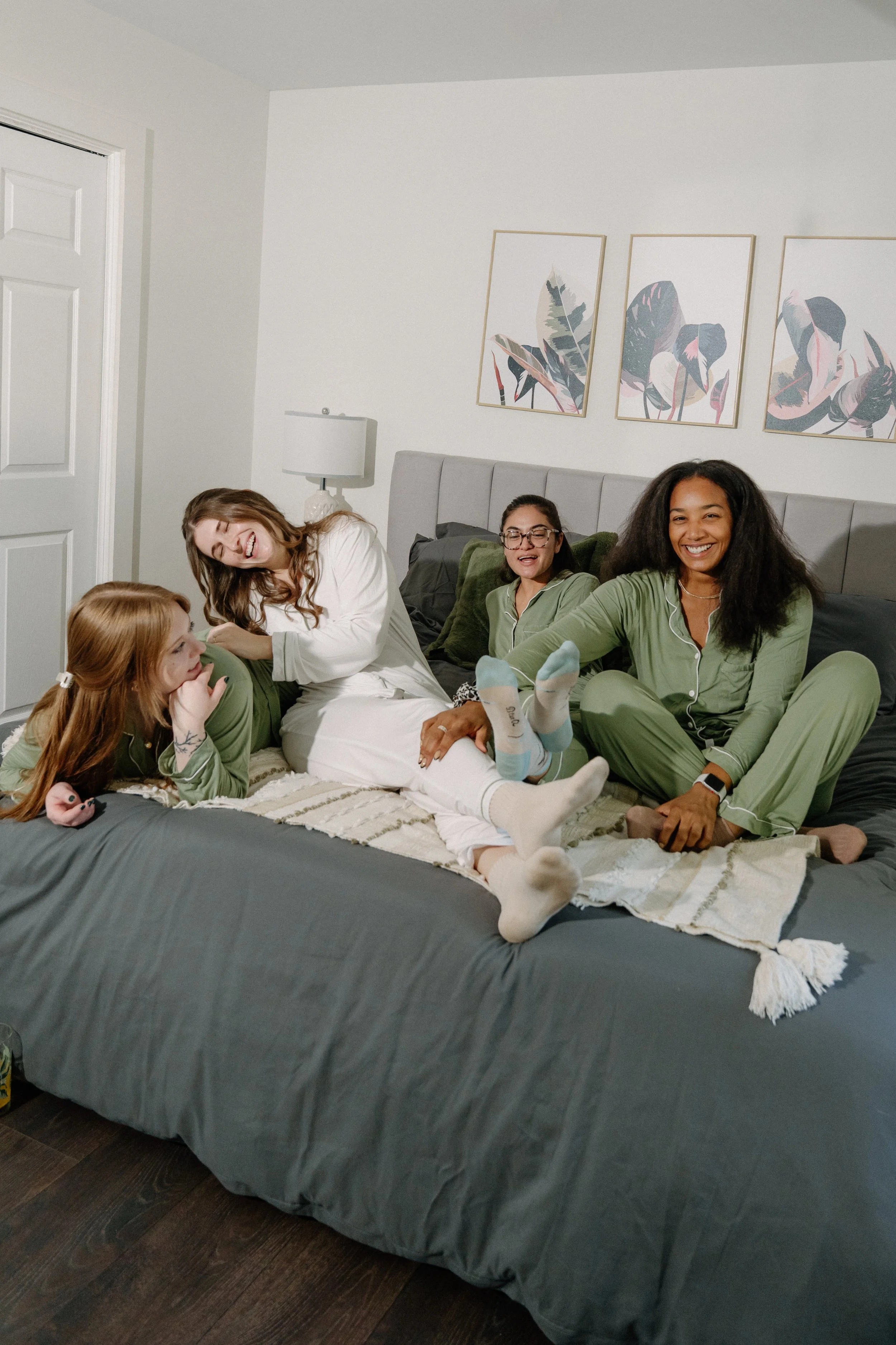 Four women and a girl in pajamas relaxing on a bed, laughing and smiling together in a modern bedroom with framed botanical art on the wall.