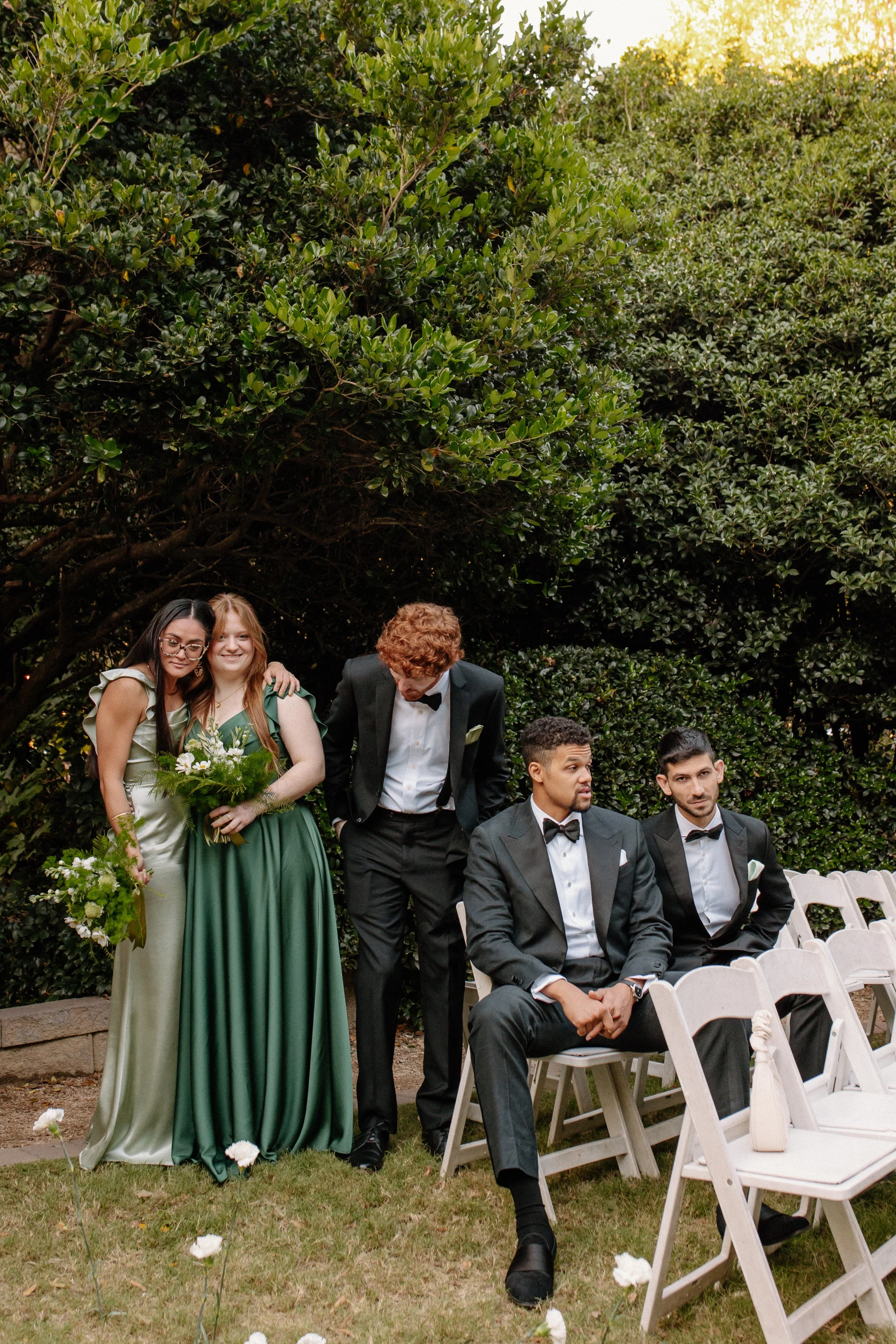 Group of five people in formal attire at an outdoor event, sitting and standing on the grass with lush greenery in the background.