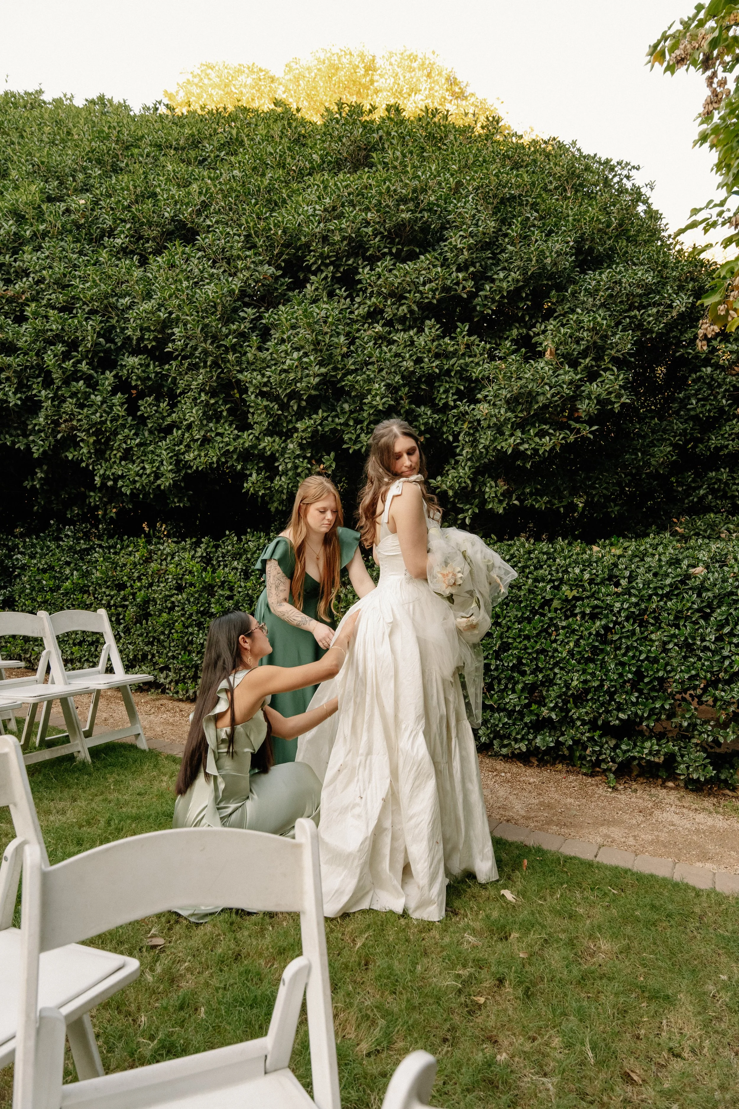 Three women in formal dresses outside, one in a white wedding gown having her dress adjusted by two others in green dresses, with chairs and greenery in the background.