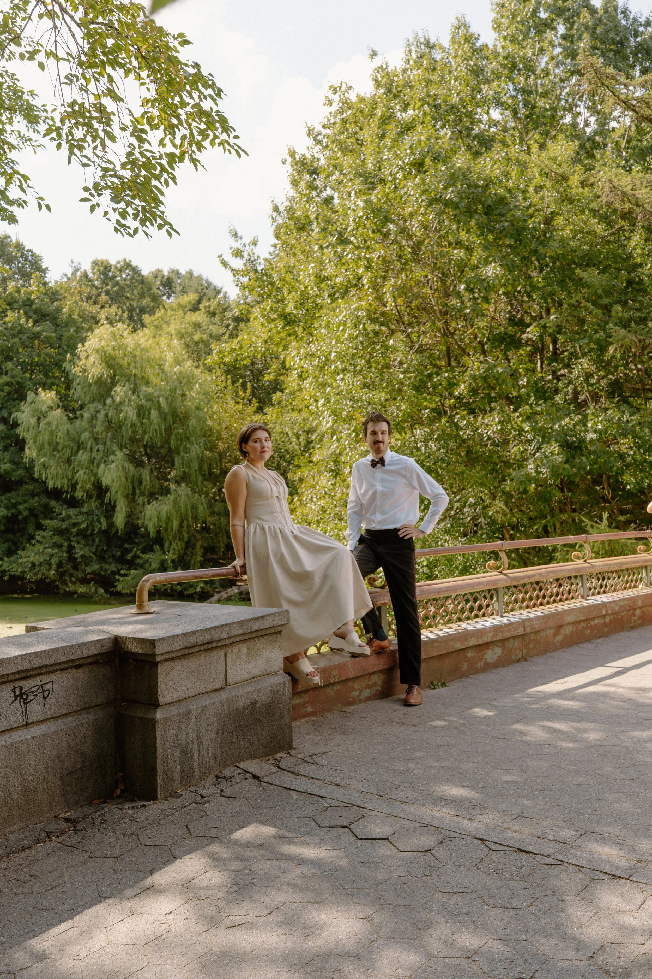 A man and woman dressed in vintage clothing posing outdoors on a stone wall and railing with trees in the background.