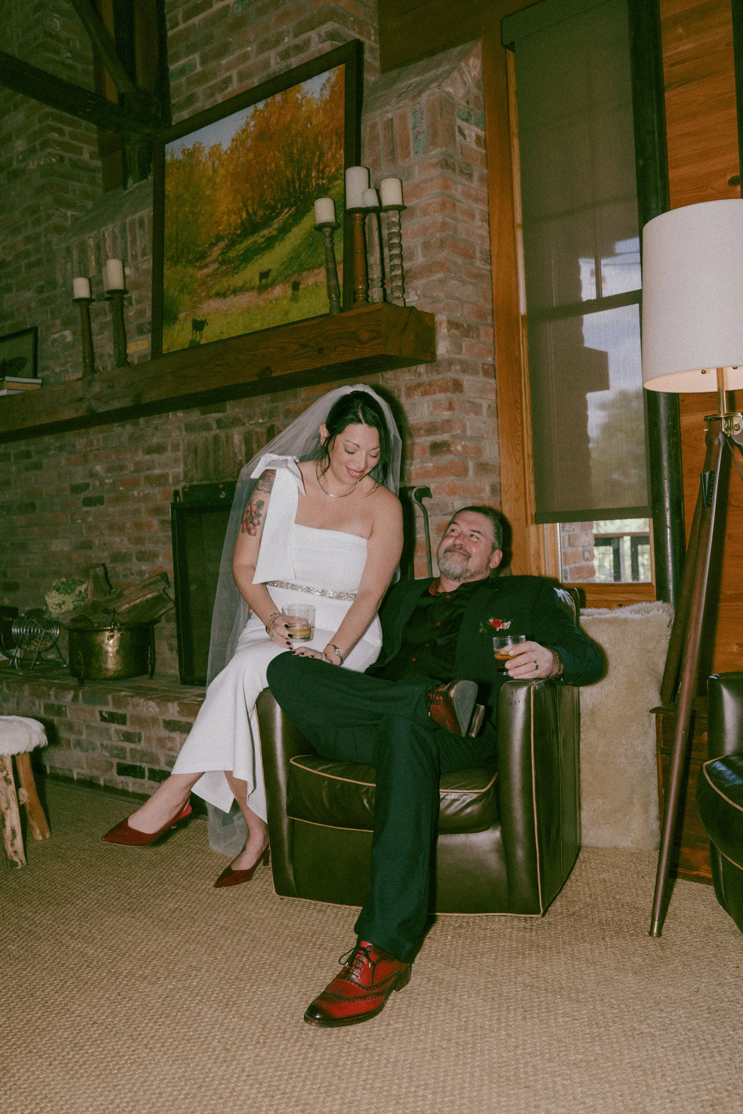 A woman in a white wedding dress with a veil and a man in a black suit sitting on a leather armchair, both smiling and holding drinks in a cozy room with a brick fireplace, art on the wall, and wooden decor.