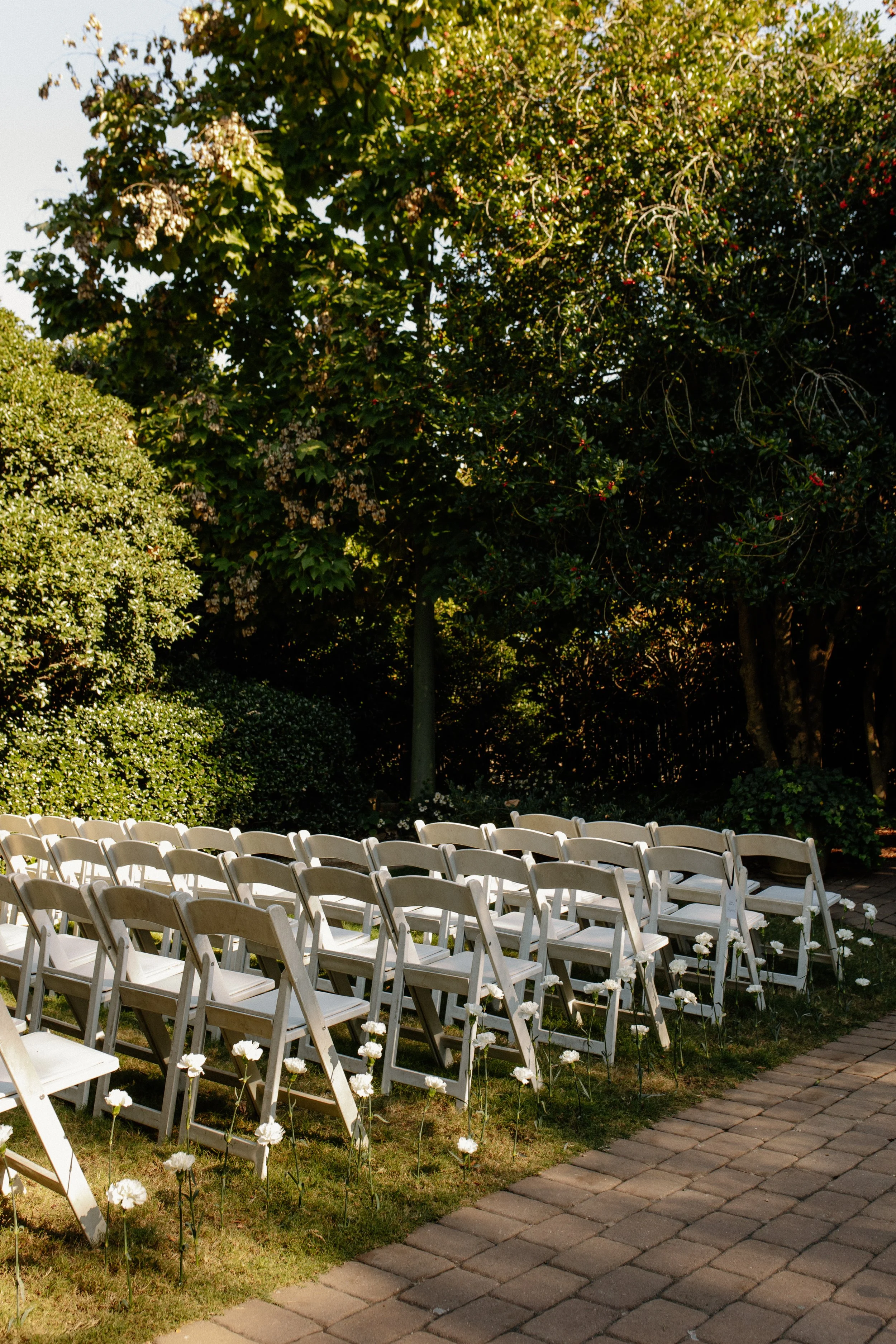 Outdoor wedding ceremony setup with white folding chairs arranged on grass next to brick pathway, decorated with white flowers, surrounded by lush green trees and bushes.