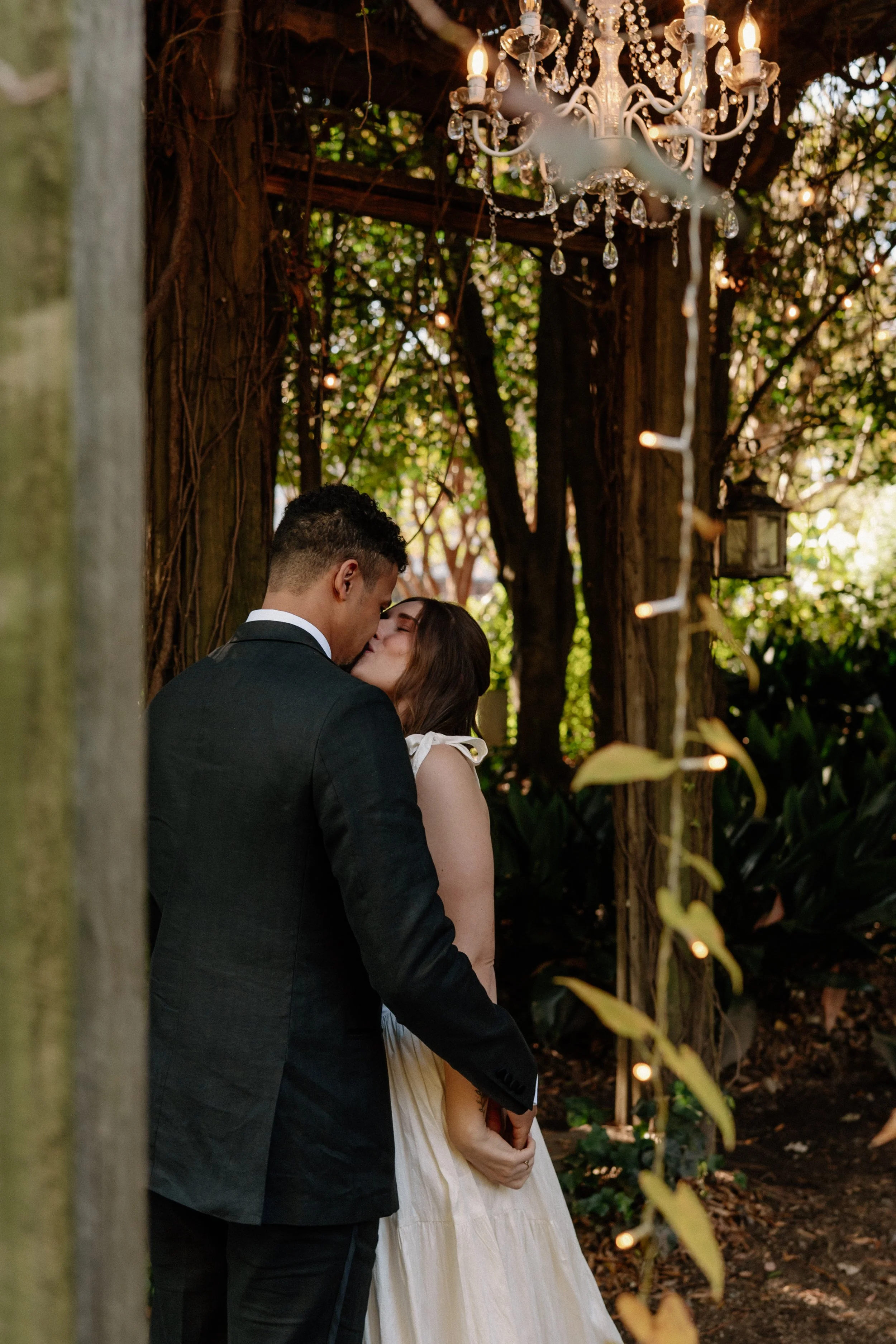 A couple shares an intimate kiss in a rustic outdoor setting decorated with a chandelier and string lights amid trees.
