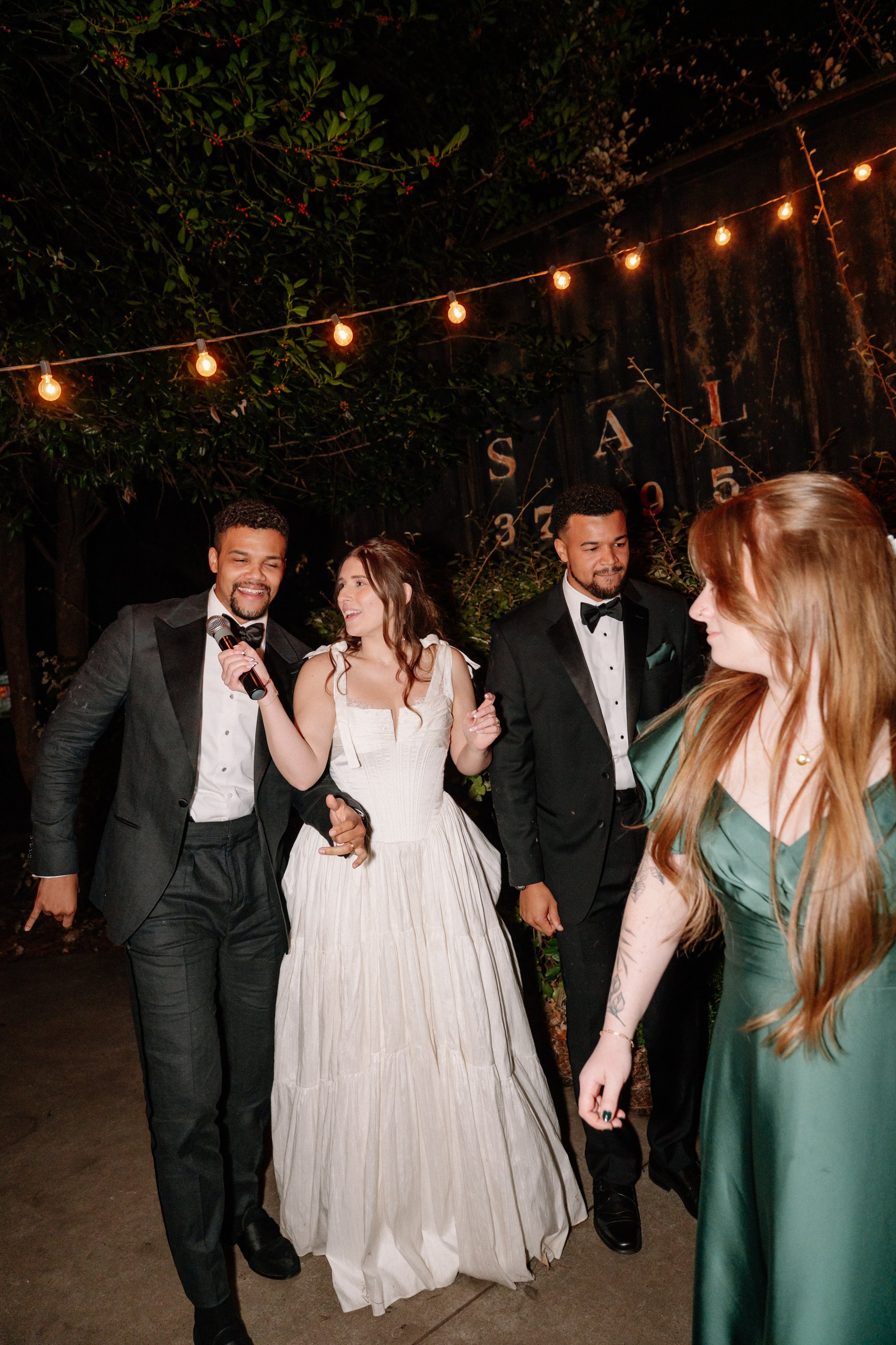 A group of five people at a wedding reception, including the bride in a white gown and four men in tuxedos, celebrating outdoors at night under string lights.