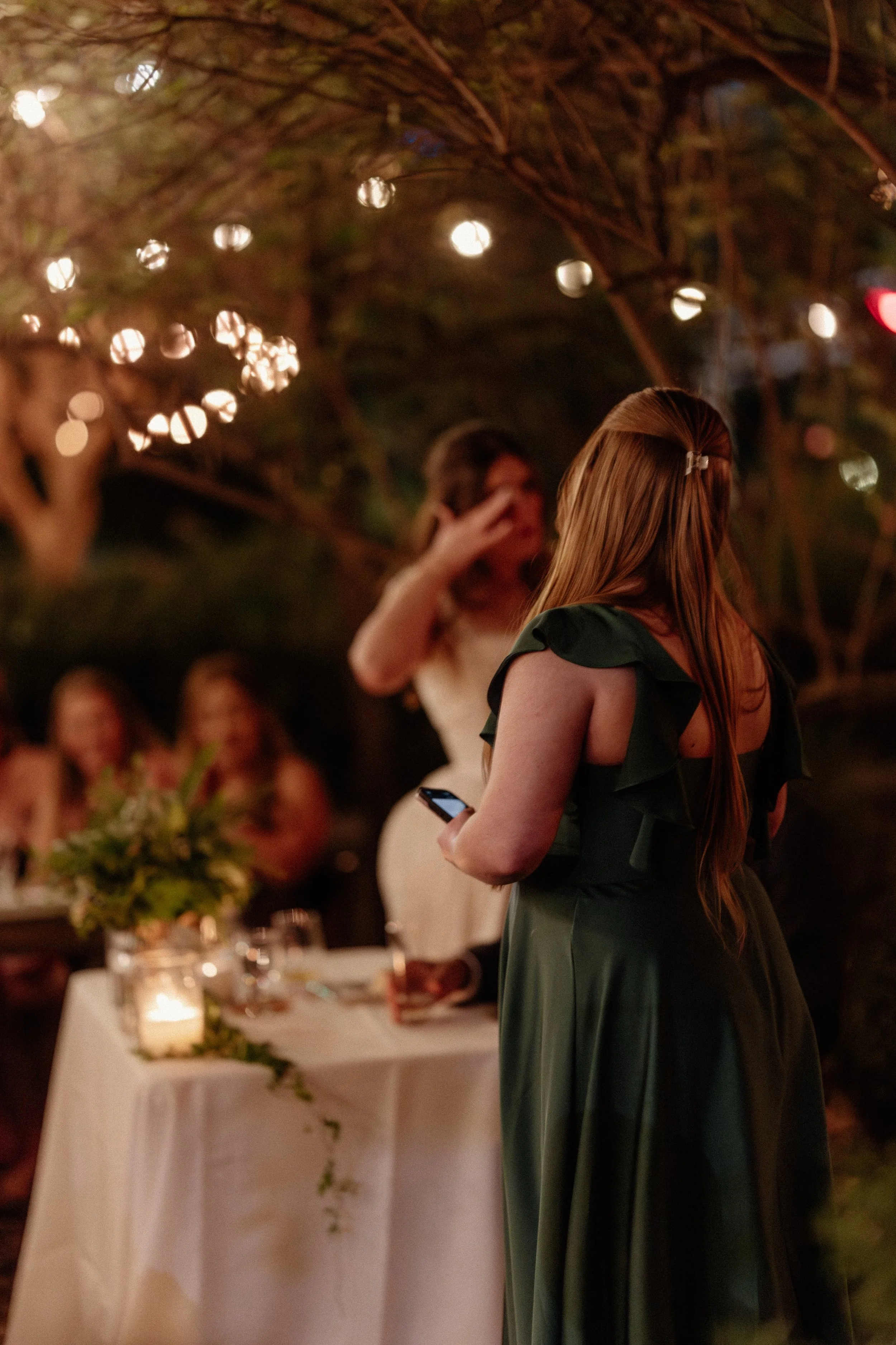 A woman with long red hair wearing a green dress is at an outdoor evening event or celebration, holding a phone in her right hand, with a table of guests and string lights above in the background.