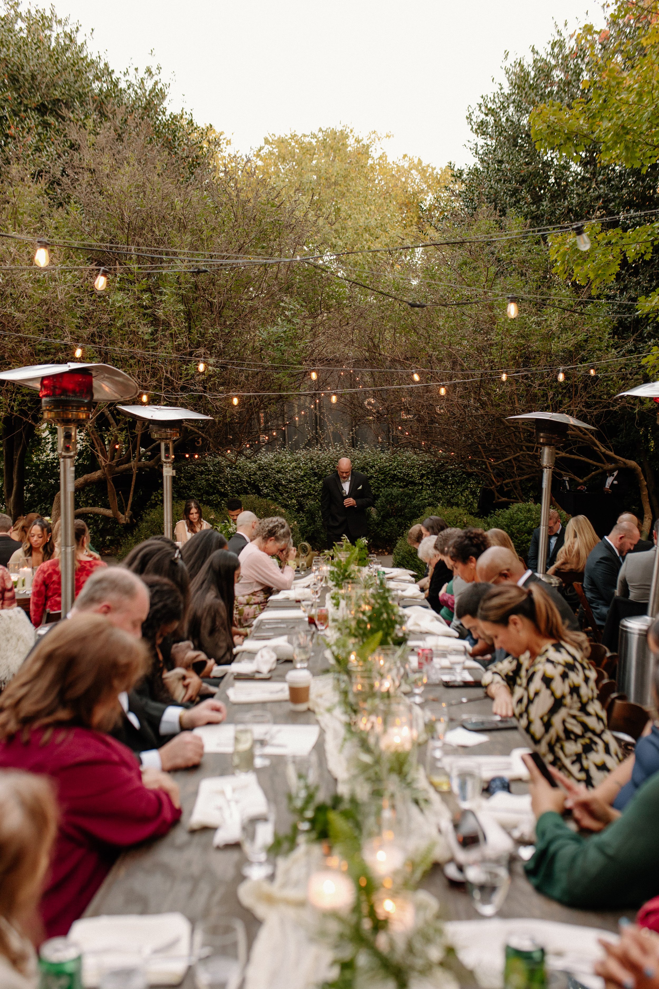 People gathered around a long outdoor dinner table, decorated with candles and greenery, under string lights and surrounded by trees.