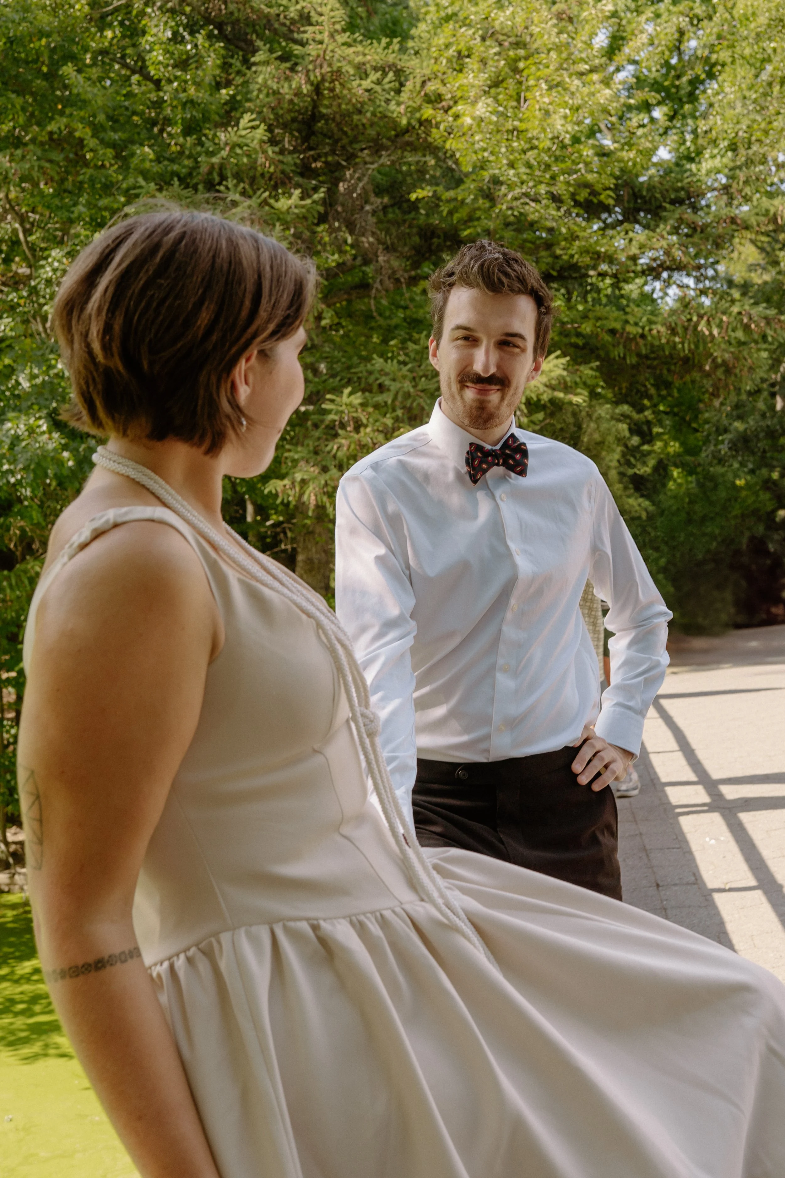 A man and woman are outdoors, engaging in conversation. The woman is wearing a sleeveless, light-colored dress and a pearl necklace. The man is dressed in a white shirt with a patterned bow tie and dark pants. They are standing near trees and a sidew