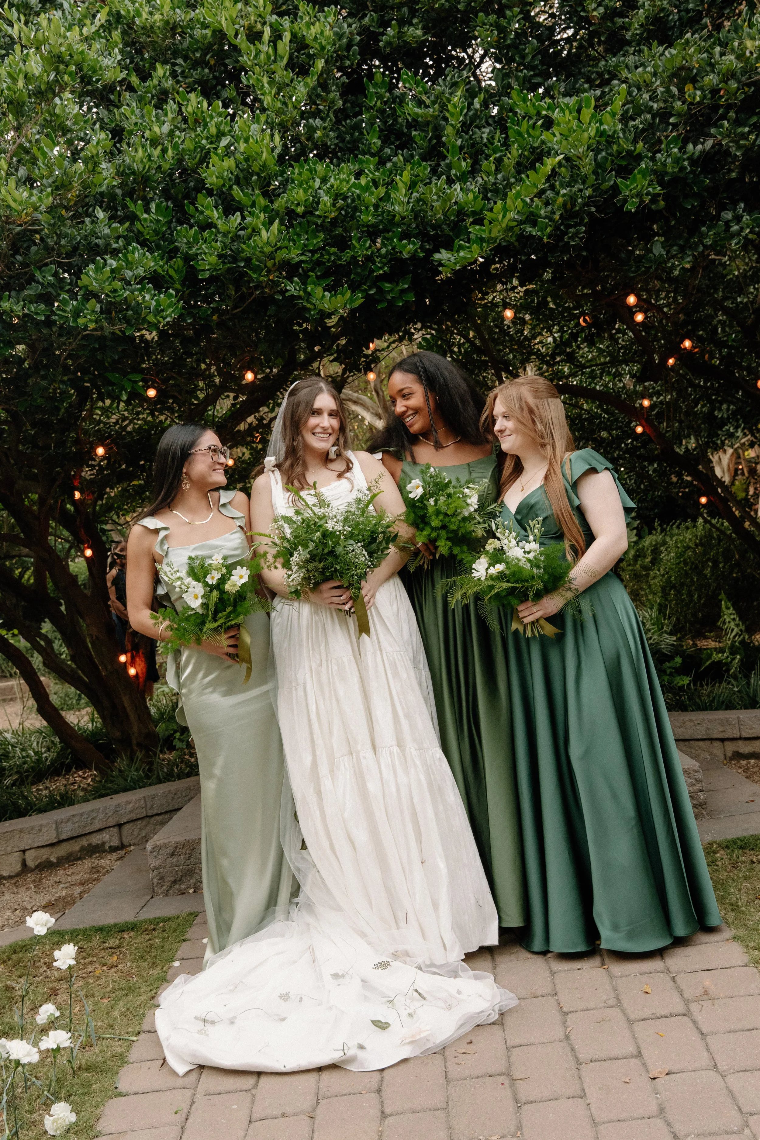 Four women in wedding attire holding bouquets of white flowers and greenery, standing outdoors under a large lush green tree with string lights, celebrating a wedding.