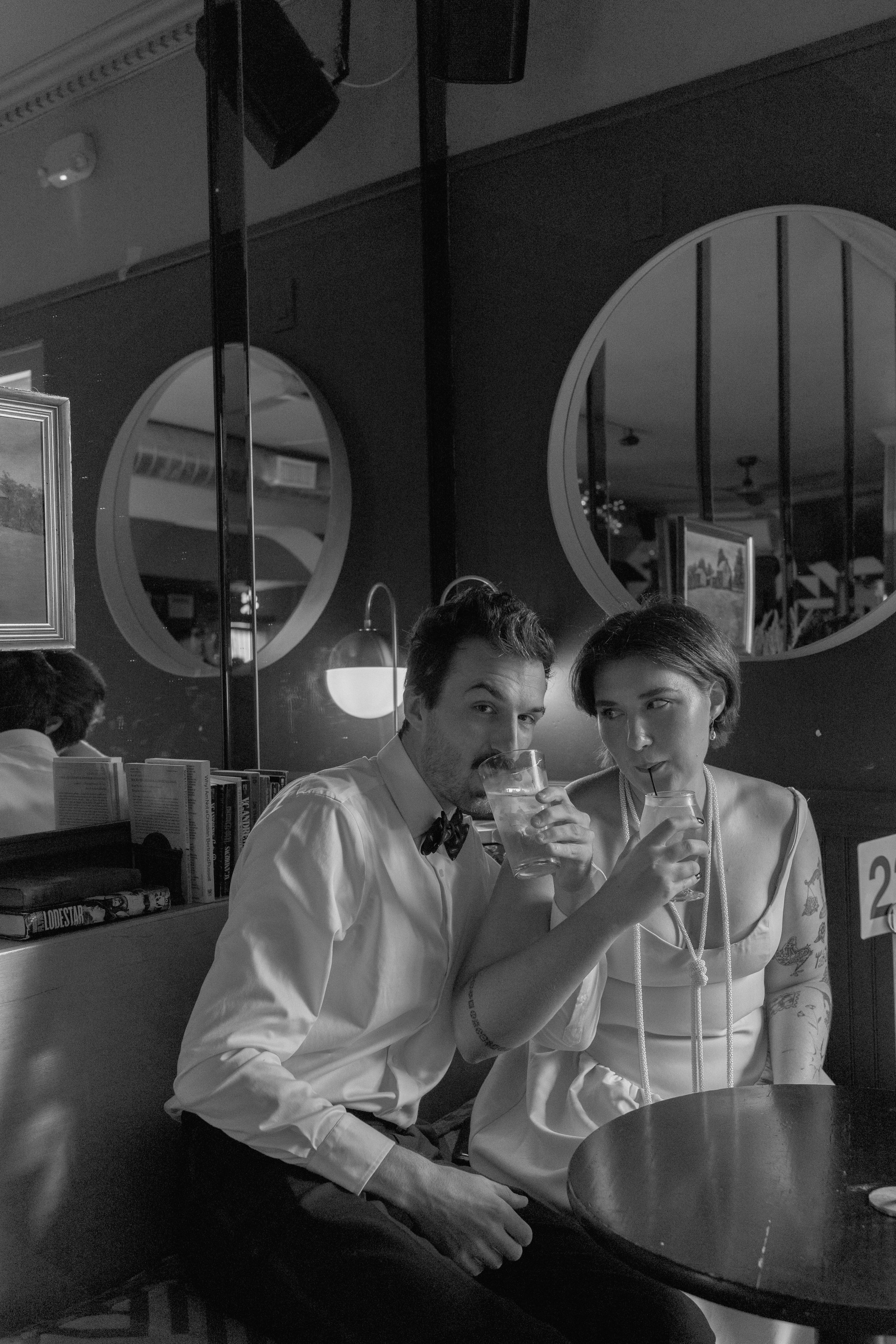 A black and white photo of a man and woman sitting at a table in a restaurant or bar, drinking from glasses with straws and looking at the camera. The man is wearing a white shirt and a bow tie, the woman has short hair and tattoos on her arms.