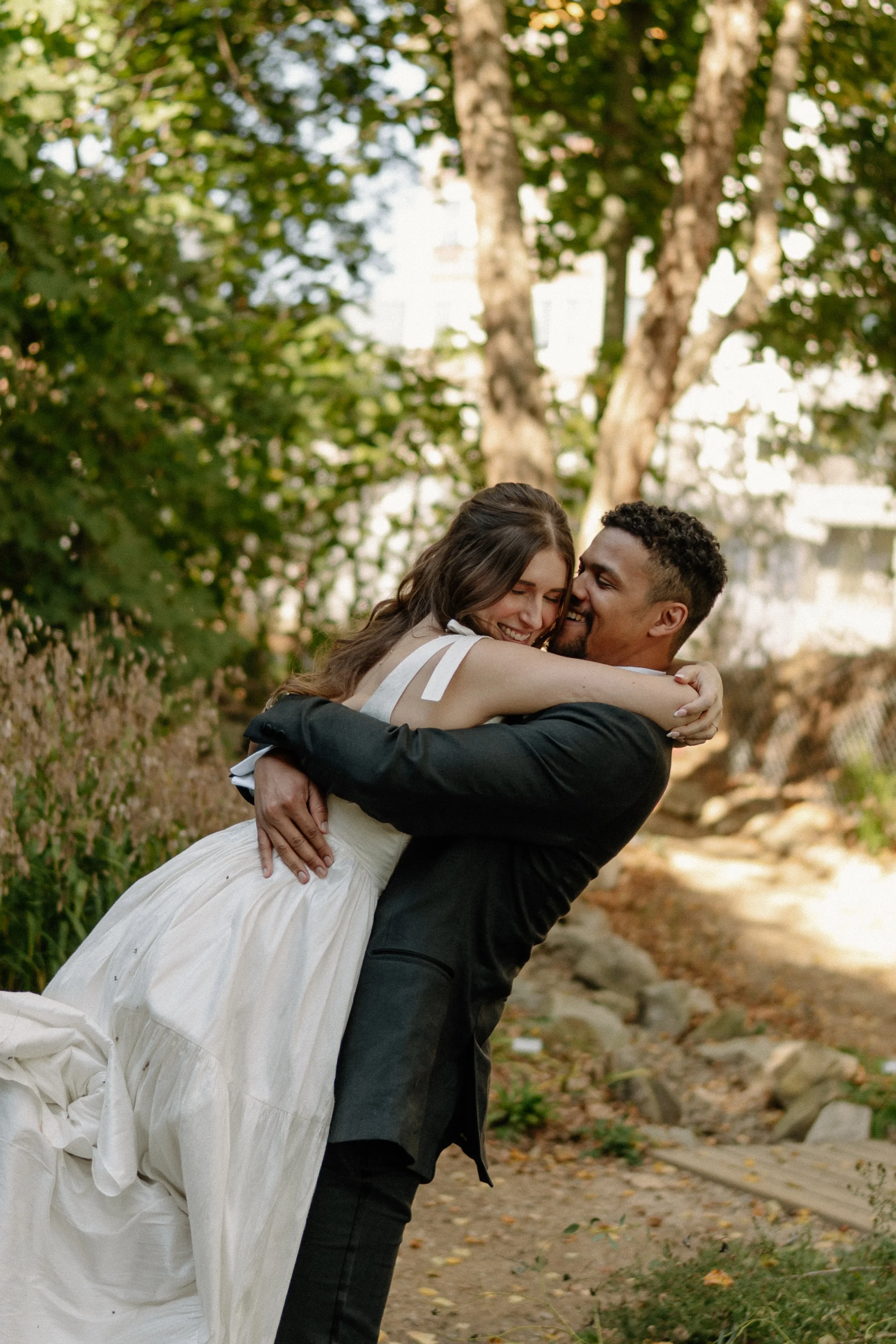 A man in a black suit holding a woman in a white dress and smiling, outdoors with trees and rocks in the background.