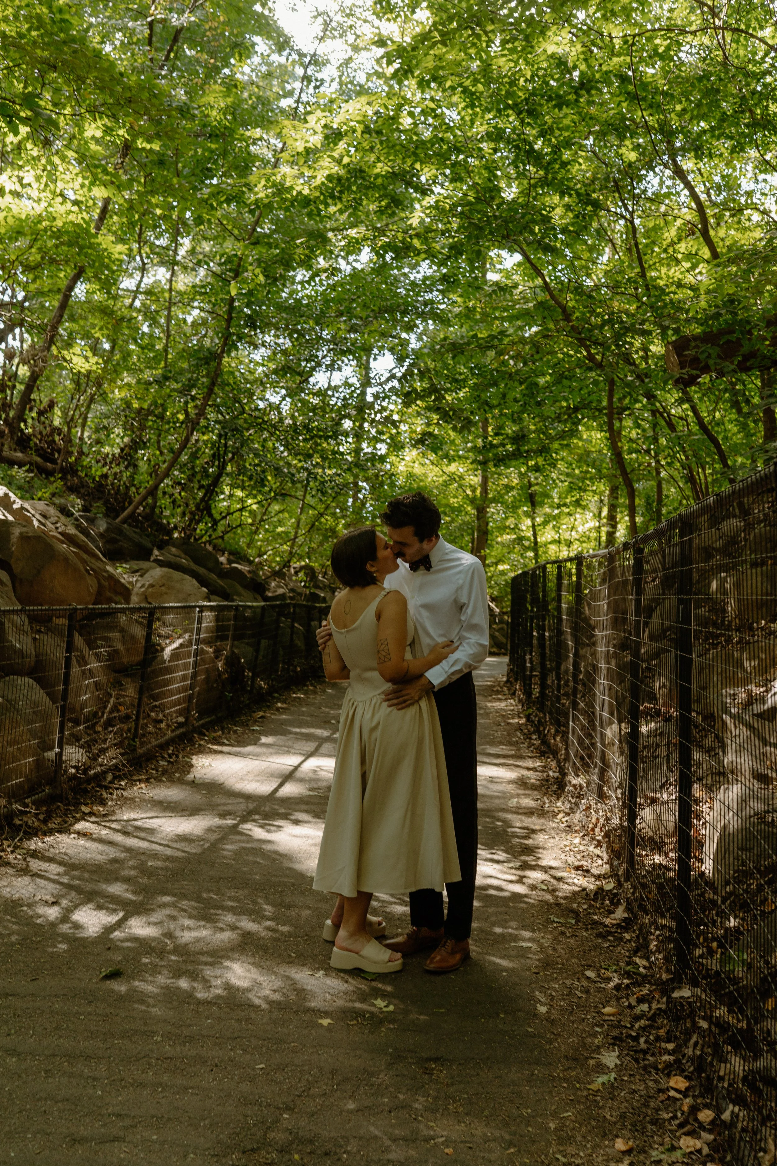 A couple romantically embracing on a wooded pathway surrounded by trees, sunlight filtering through leaves.