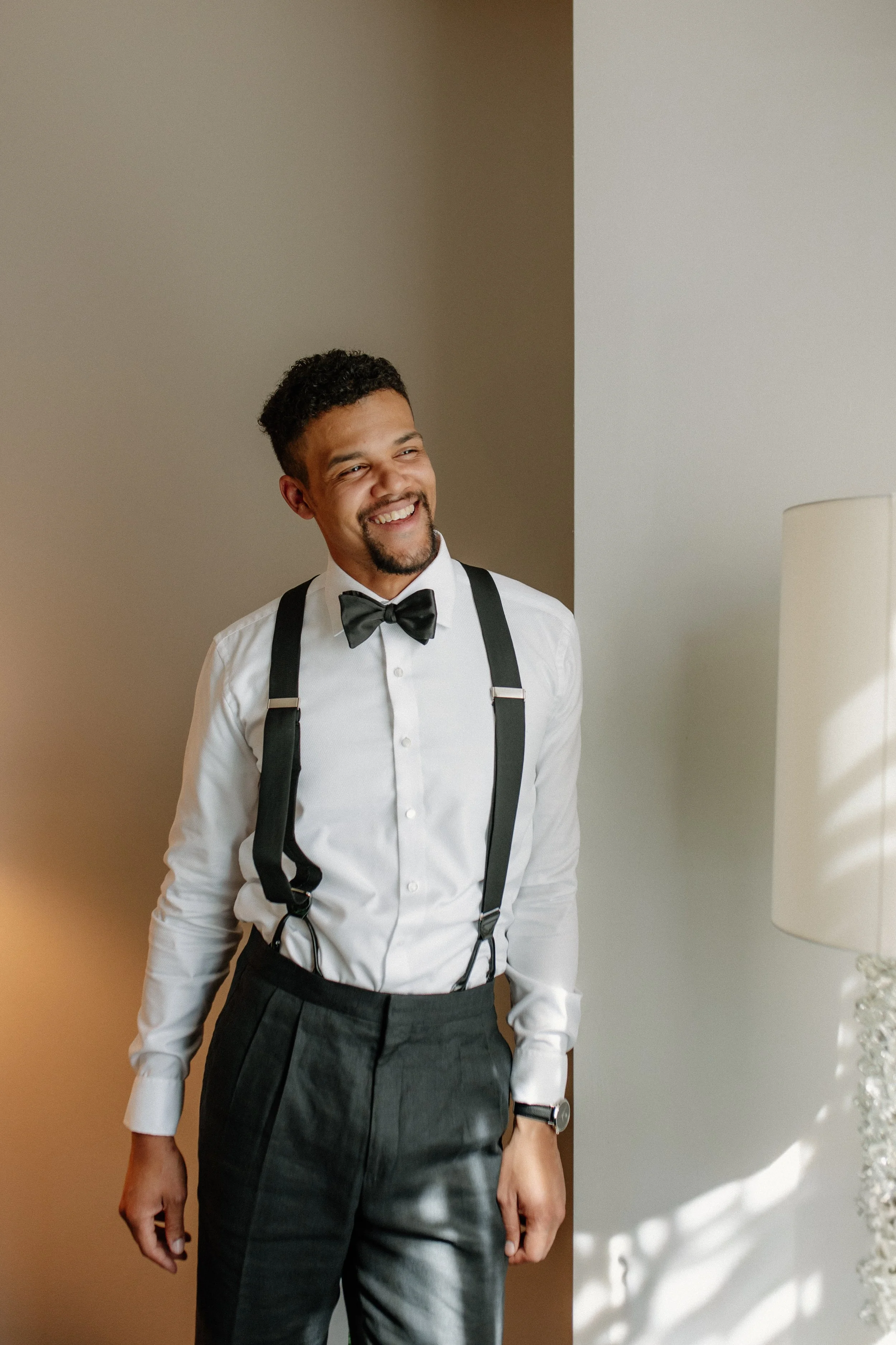 Young man dressed in formal attire with a white shirt, black bow tie, suspenders, and gray pants, smiling and standing indoors next to a lamp.