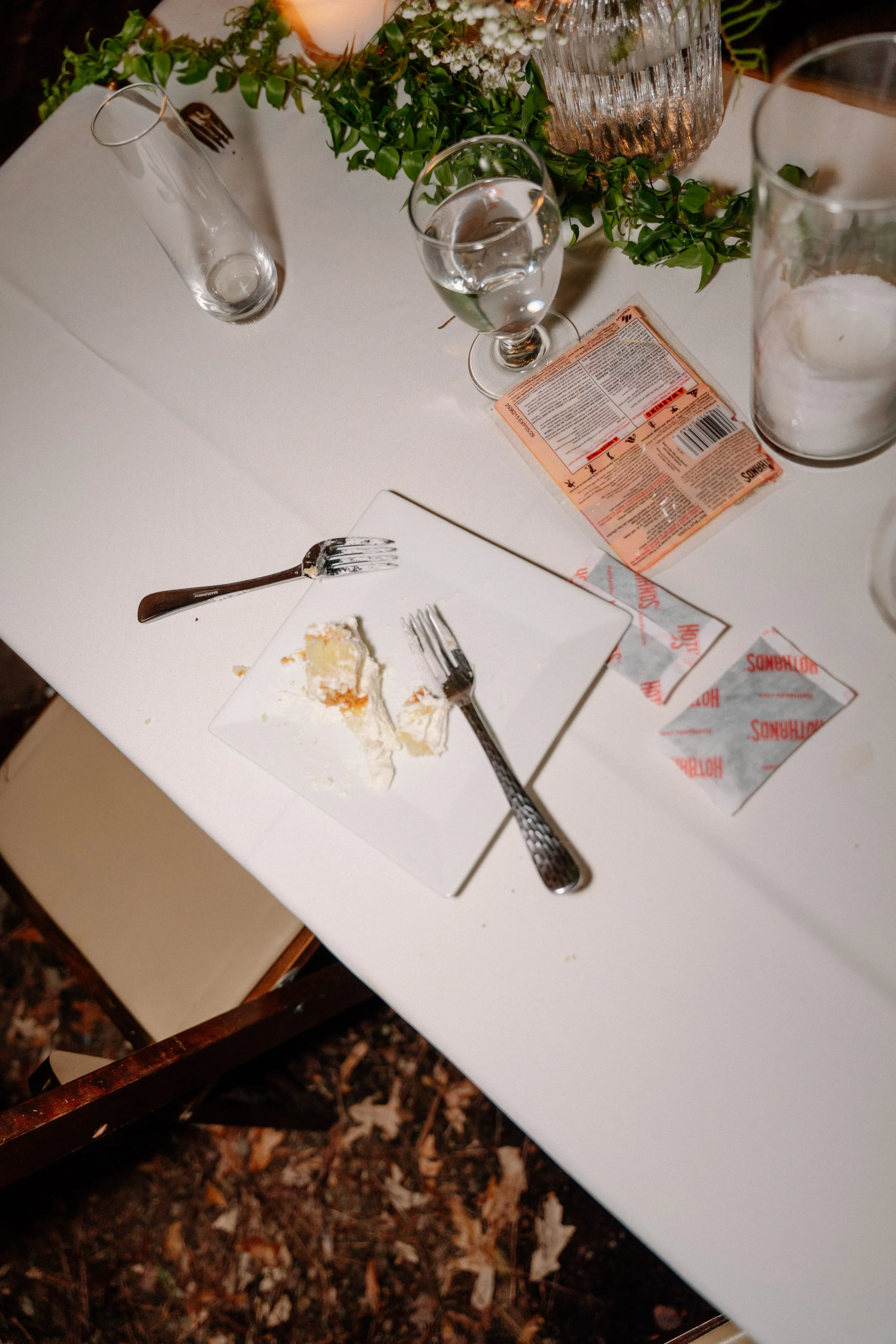 Empty dessert plate with cake crumbs and two forks on a white table, surrounded by water glasses, a candle, a floral centerpiece, and condiments.