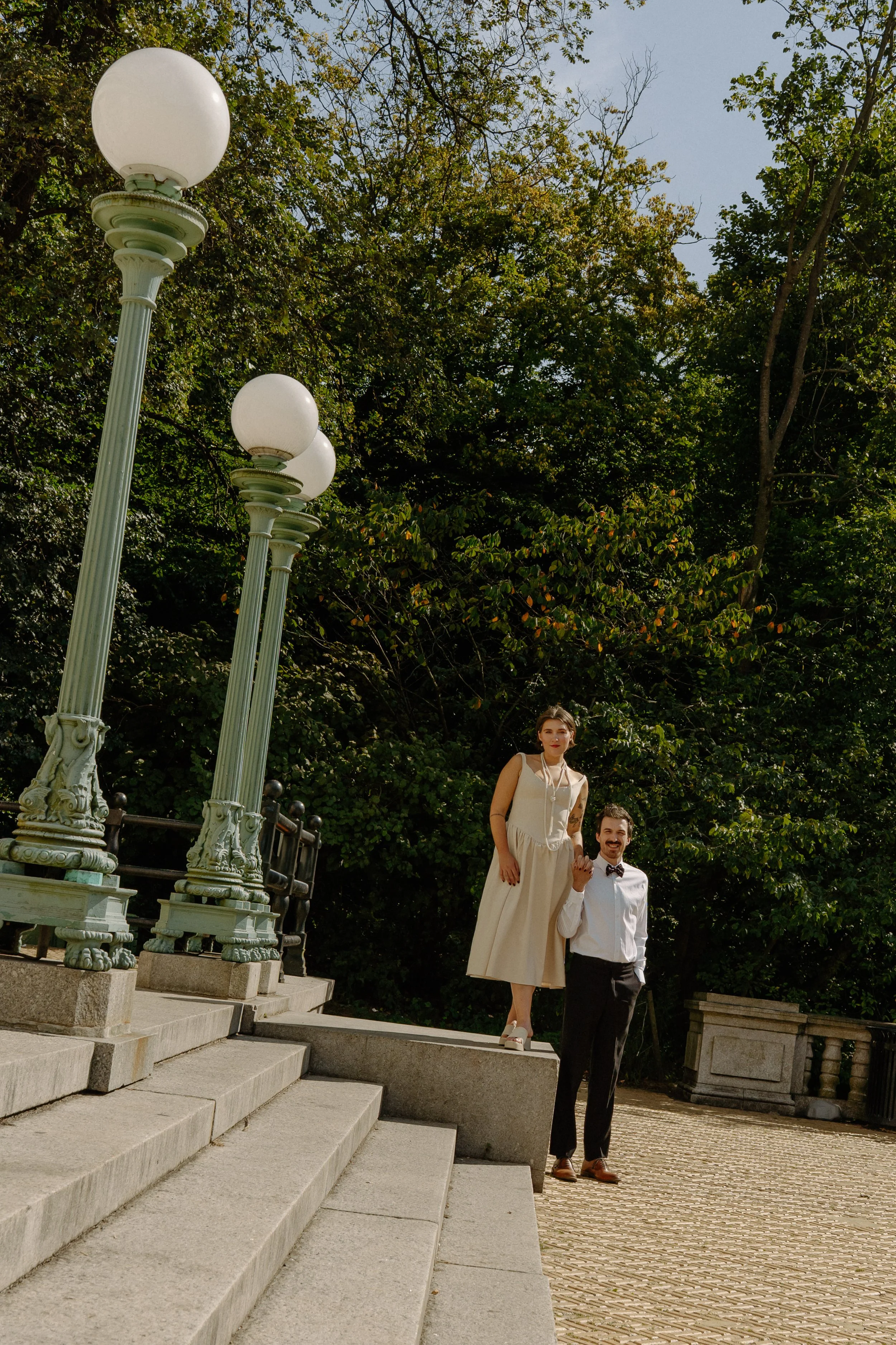 A man in a white shirt, black pants, and a bow tie holding a woman in a cream-colored dress and white shoes on a stone ledge outdoors surrounded by trees and vintage street lamps.