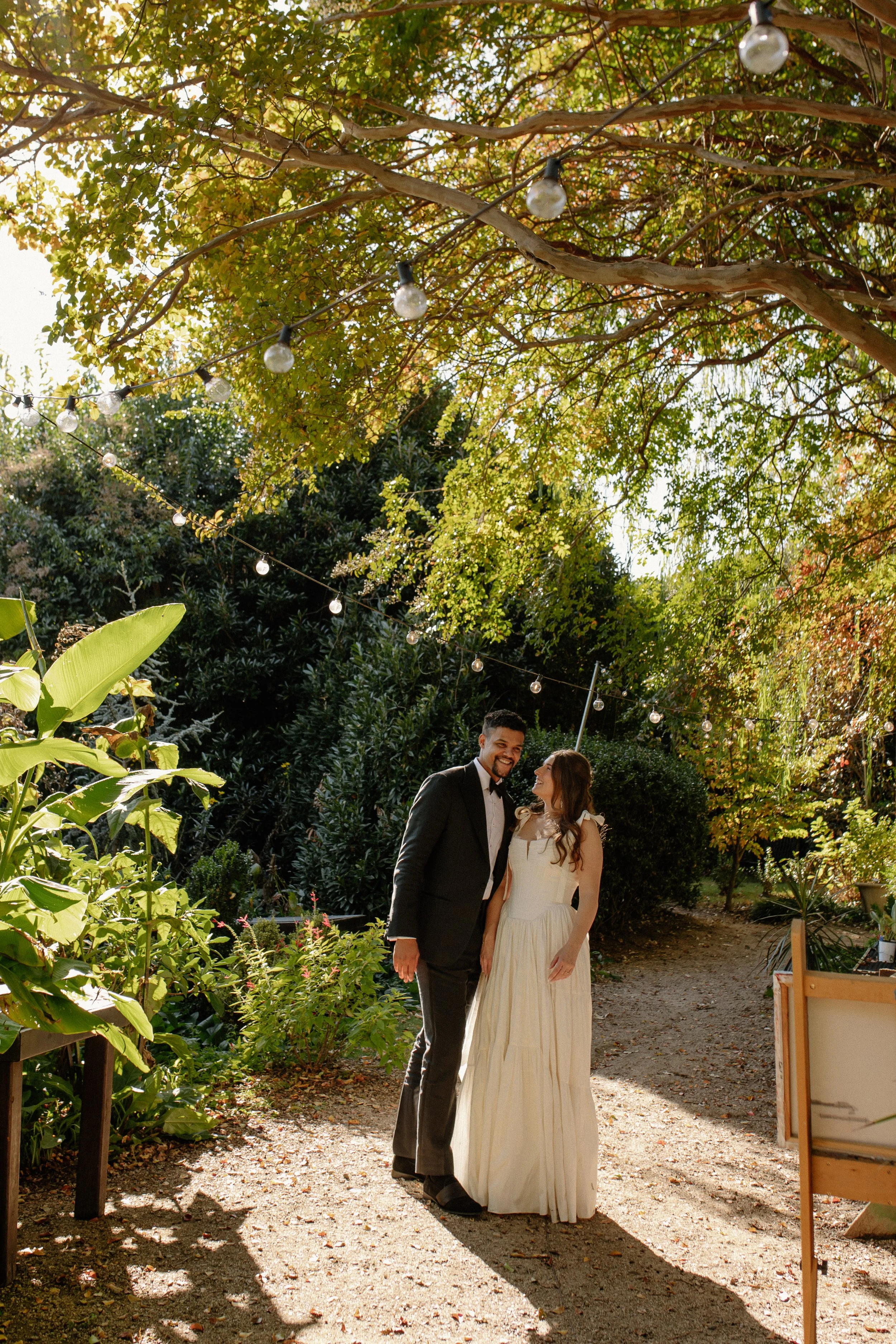 A couple dressed in formal wedding attire celebrating outdoors under string lights and trees, smiling at each other.
