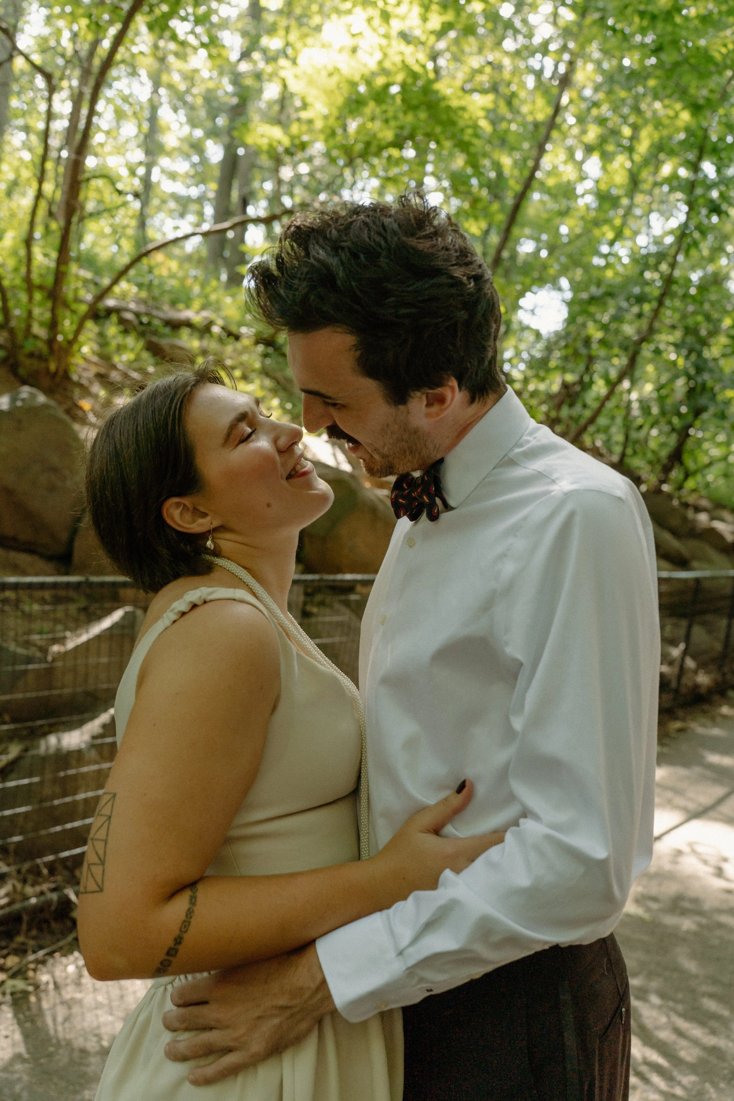 A couple with light skin and dark hair smiling and looking into each other's eyes while standing close together outdoors in a wooded area.
